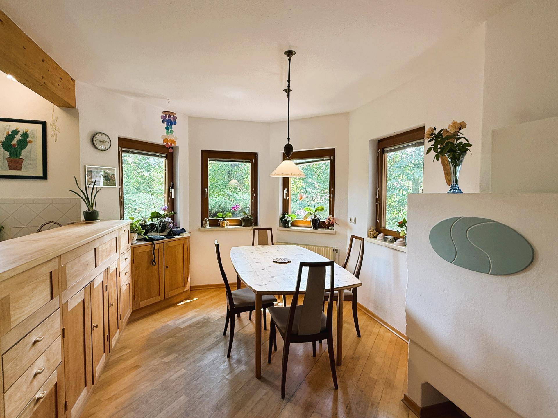 Essbereich mit Holztisch und Stühlen, umgeben von Fenstern mit Blick ins Grüne.