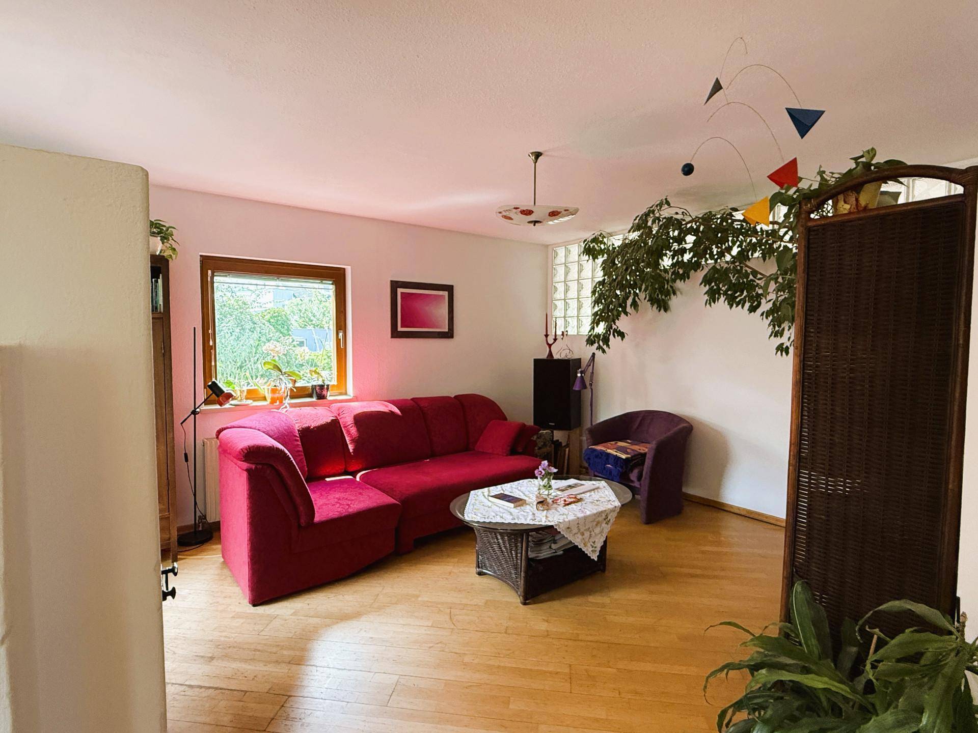Gemütliches Wohnzimmer mit rotem Sofa, Holzboden und Blick ins Grüne durch das Fenster.