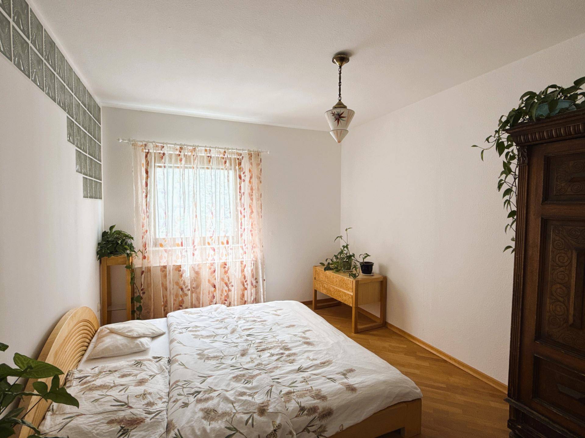 Helles Schlafzimmer mit Holzbett, floraler Bettwäsche und Fenster mit Blick ins Grüne.