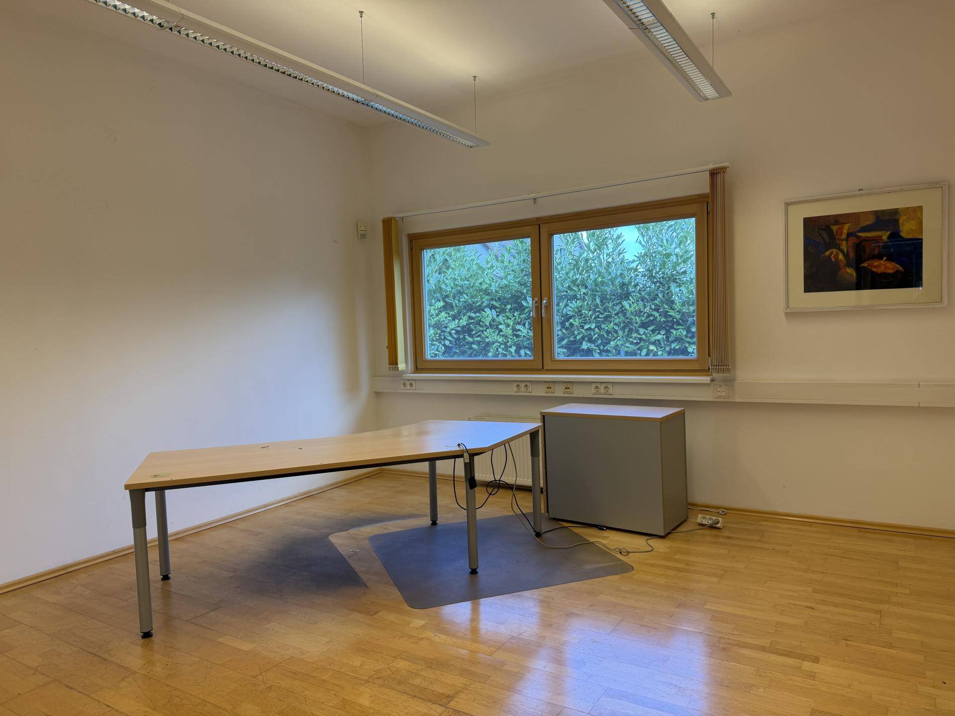 Leeres Büro mit Holzboden, großem Fenster und einem Schreibtisch in L-Form.