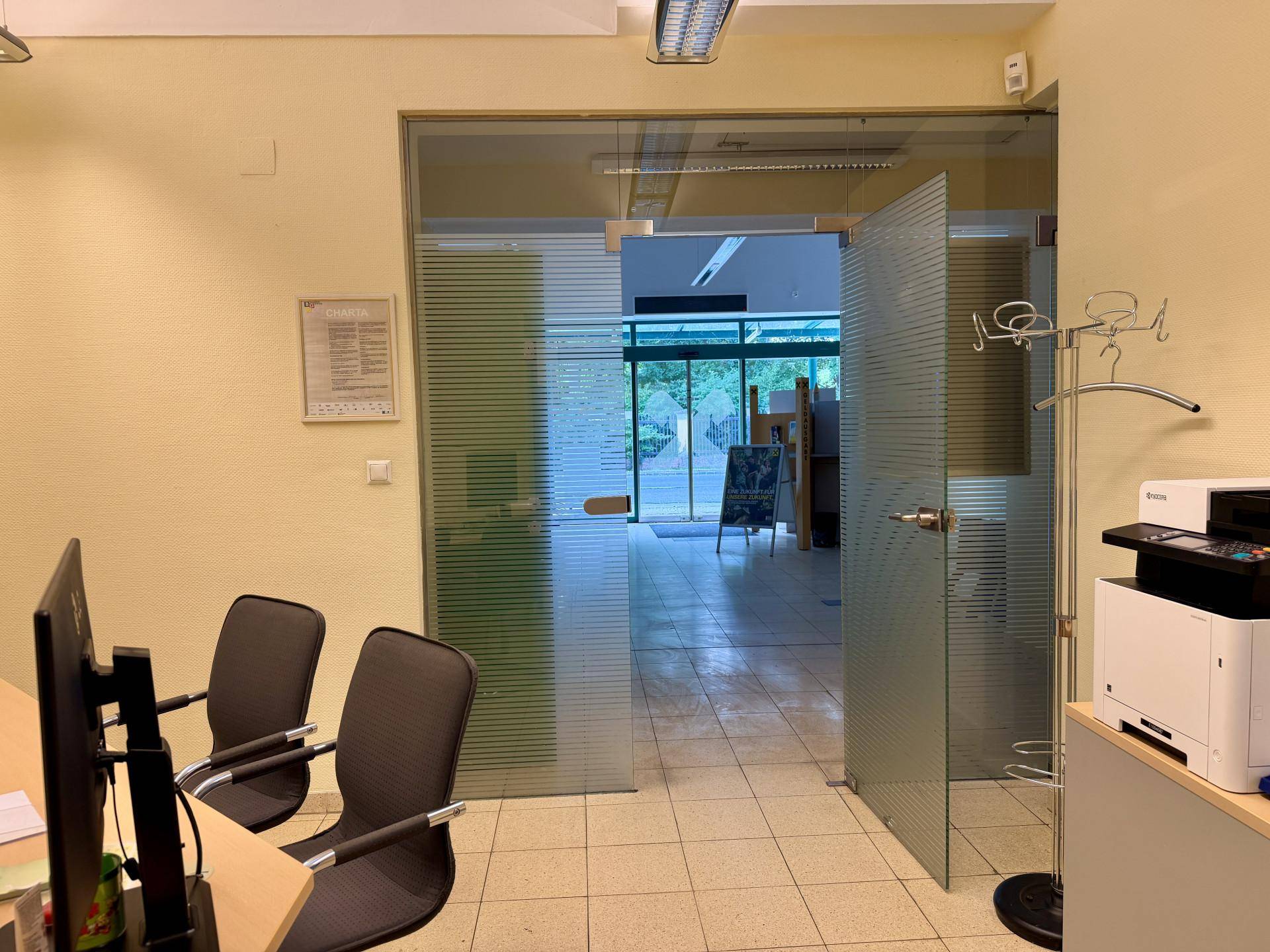 Blick aus einem Büro durch eine Glastür in den Empfangsbereich mit Fliesenboden.