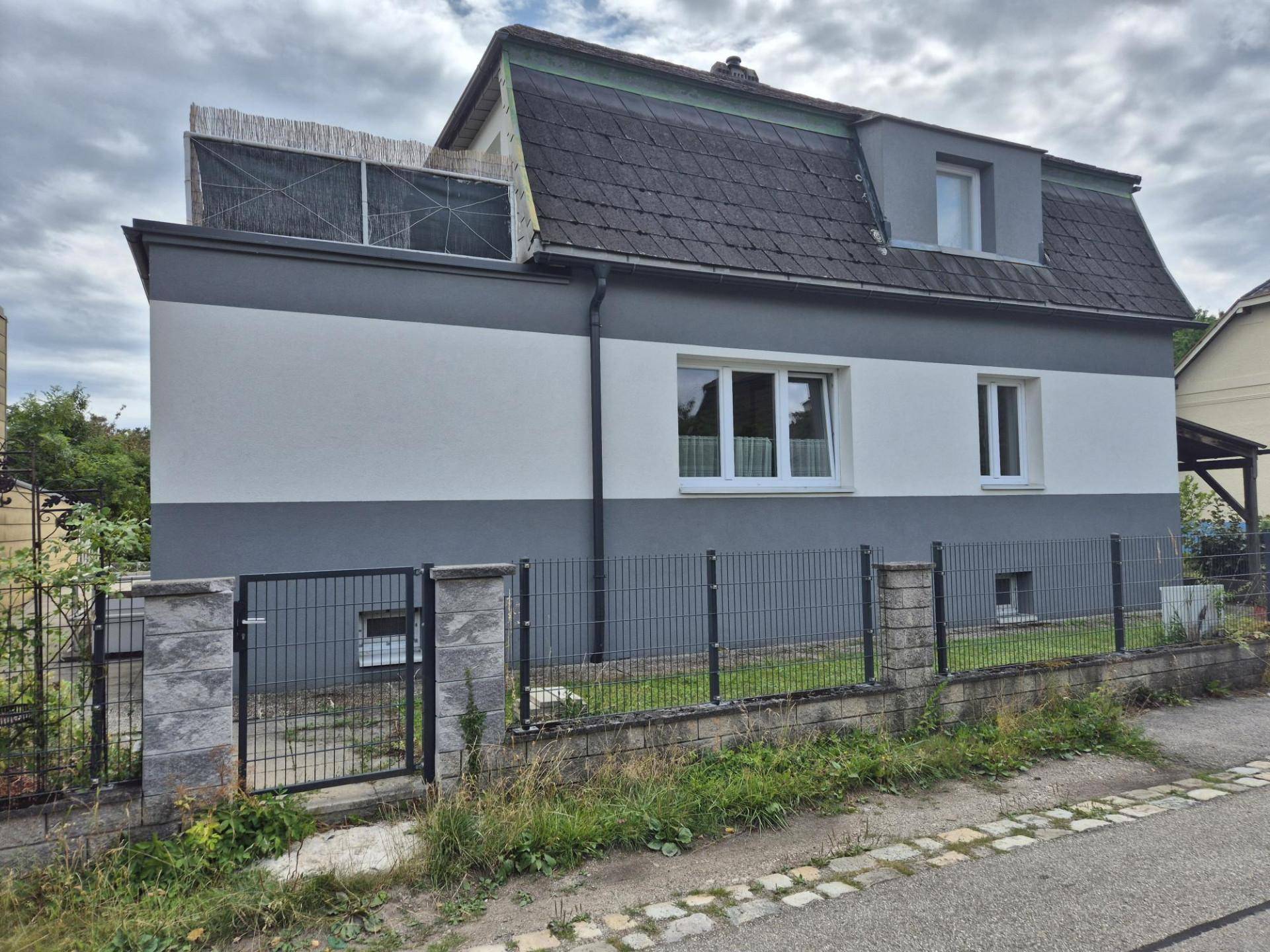 Seitenansicht des Hauses mit grauer und weißer Fassade unter bewölktem Himmel.
