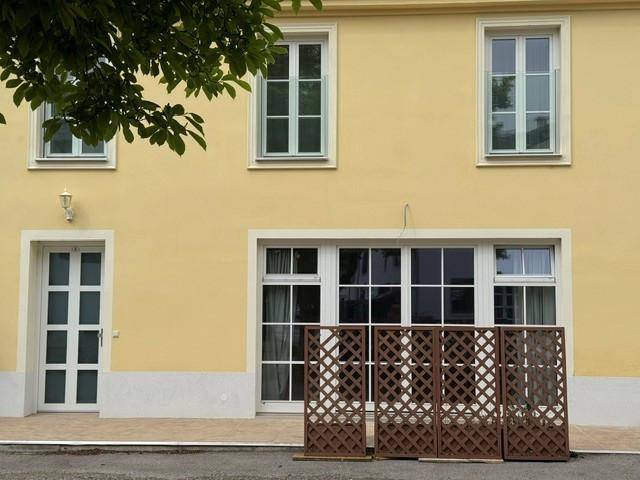 Fassade eines gelben Gebäudes mit Fenstern und einer Eingangstür, umgeben von Grün.