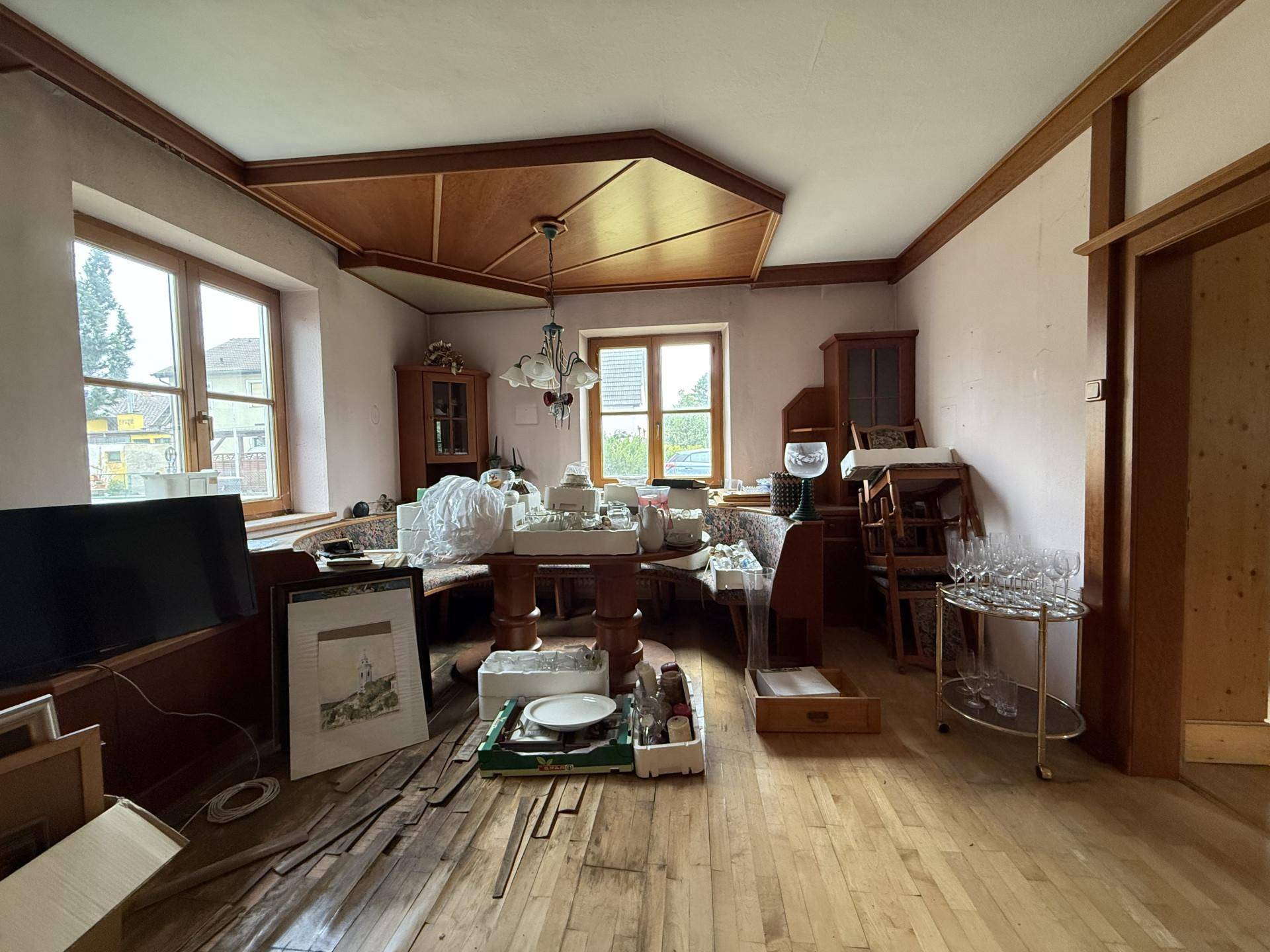 Ein unaufgeräumtes Wohnzimmer mit Holzboden, Holzvertäfelung und Blick auf den Garten durch die Fenster.