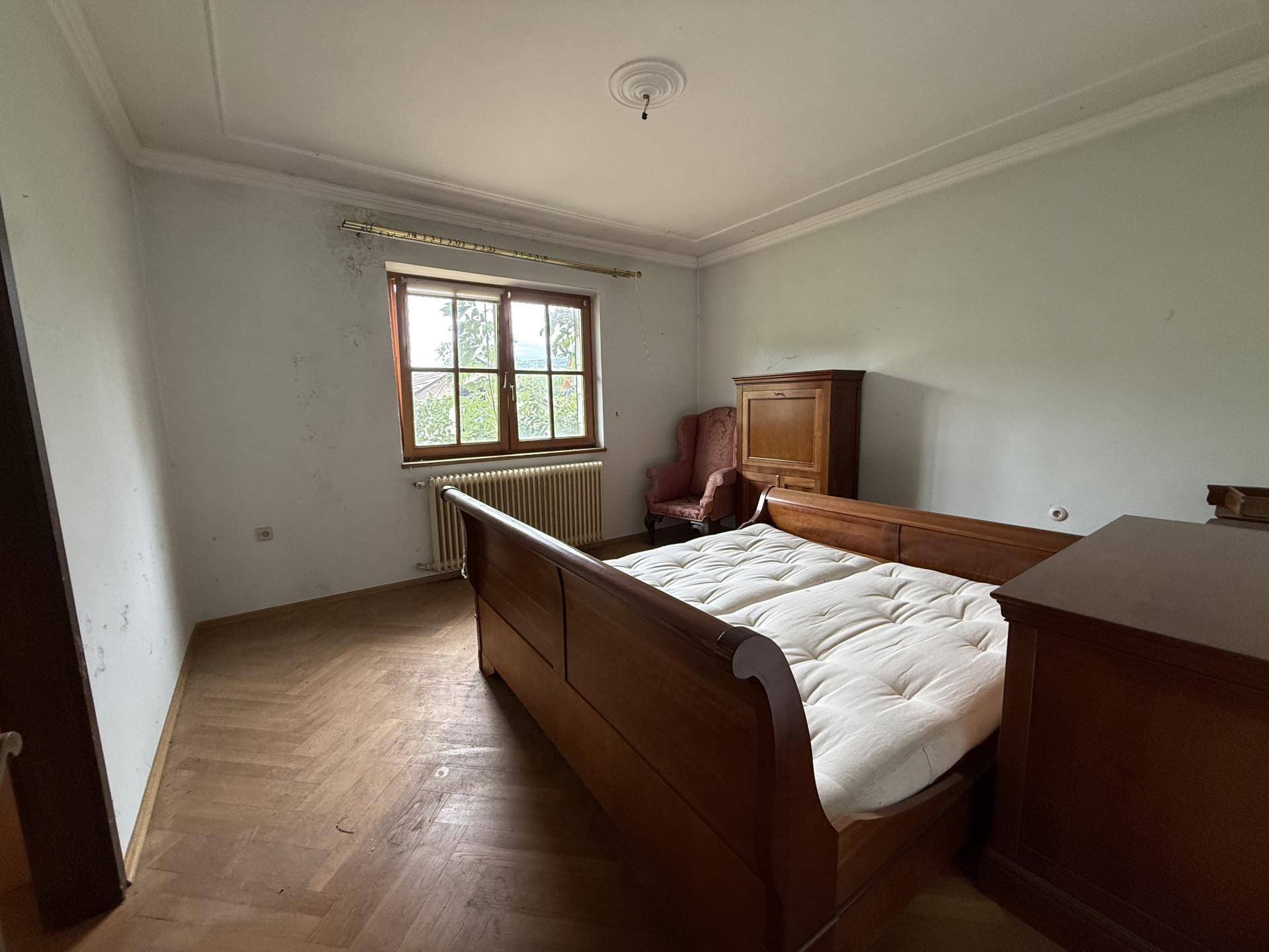 Ein Schlafzimmer mit Holzbett, Fischgrätparkett und einem Fenster mit Blick ins Grüne.