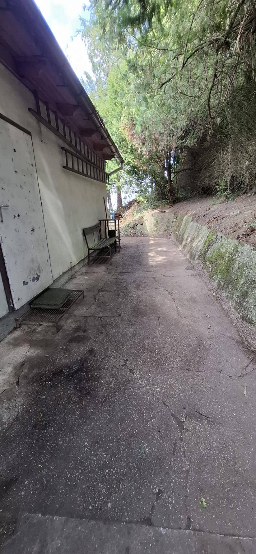 Schmaler Betonweg entlang eines Nebengebäudes mit einer Bank und Bäumen.
