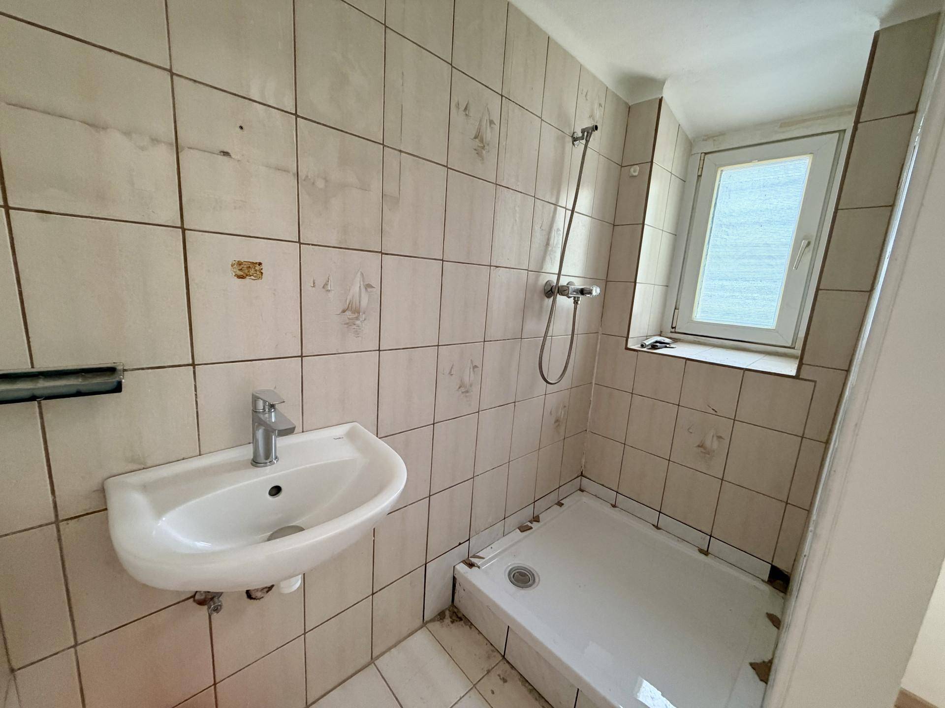 Kleines Badezimmer mit beigen Fliesen, Waschbecken und Duschbereich, ausgestattet mit einem Fenster.