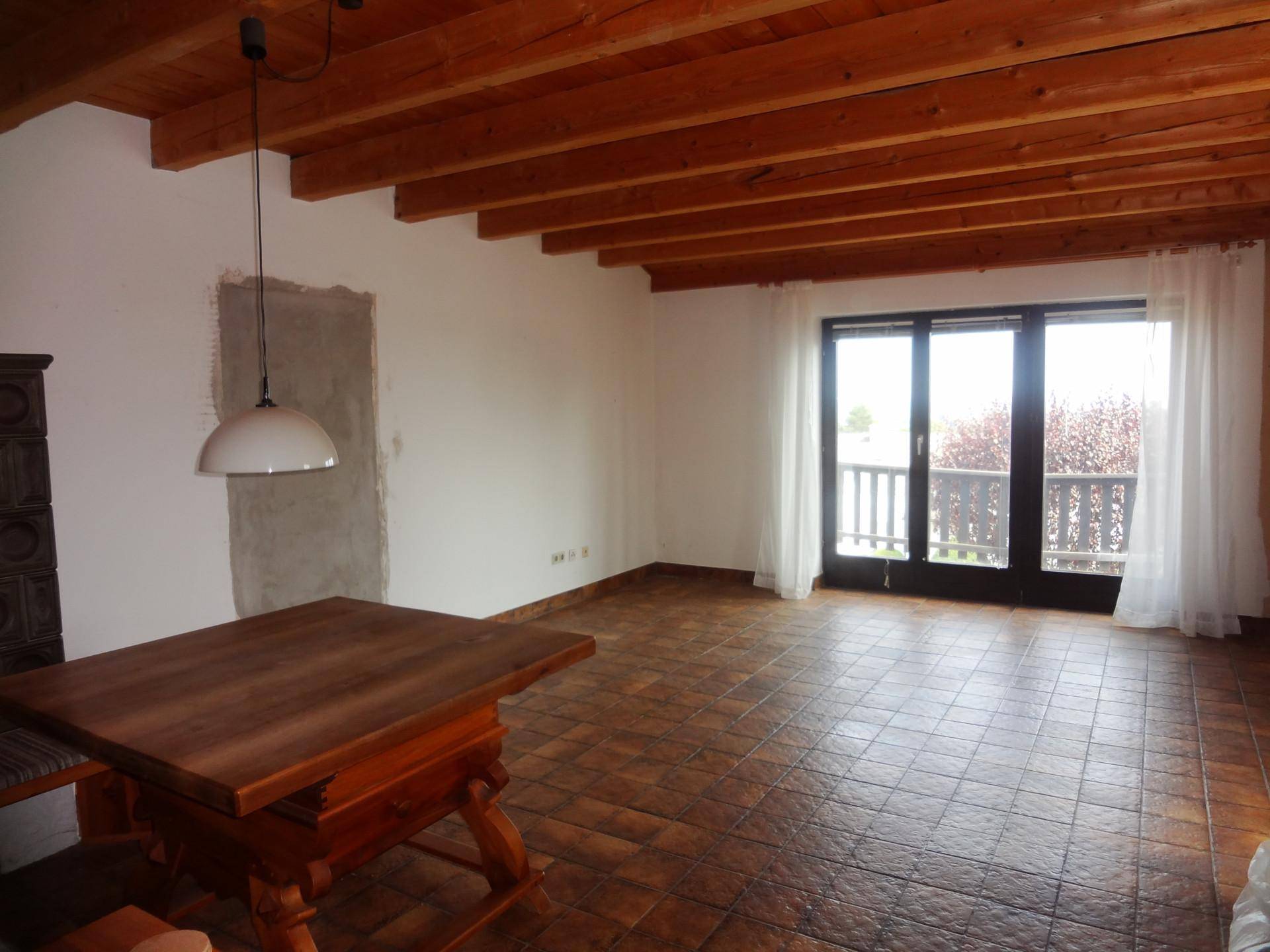 Rustikaler Wohn-Essbereich mit sichtbaren Holzbalken, Fliesenboden und Zugang zum Balkon.