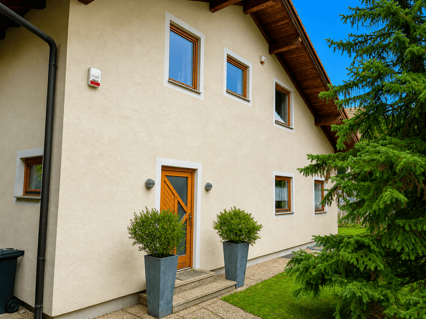 Eingangsbereich des Hauses mit beiger Fassade, Holztür und dekorativen Pflanzen in hohen Kübeln.