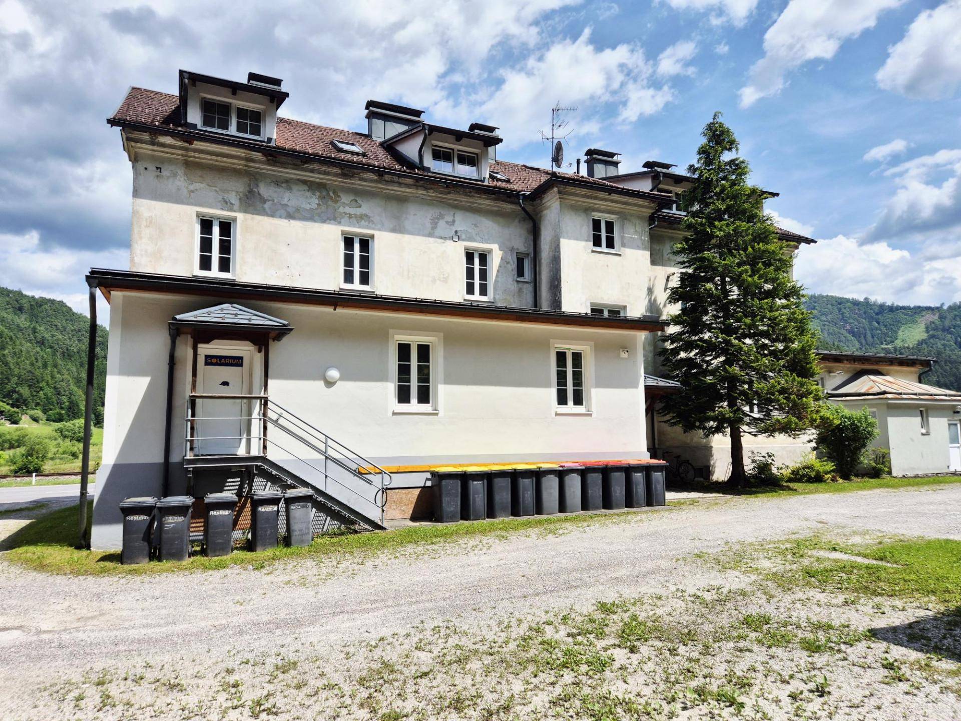 Gebäudeeingang mit Treppe und mehreren Mülltonnen, umgeben von Schotter und Grünflächen.