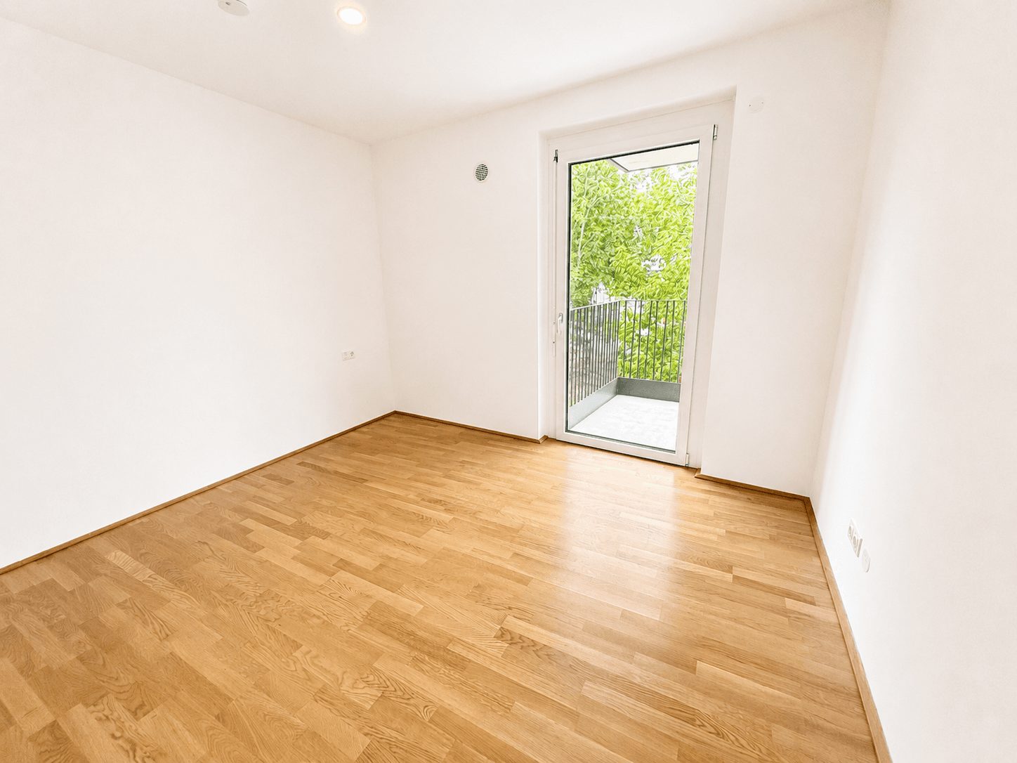 Helles Zimmer mit Holzparkettboden und direktem Zugang zu einem Balkon mit grüner Aussicht.