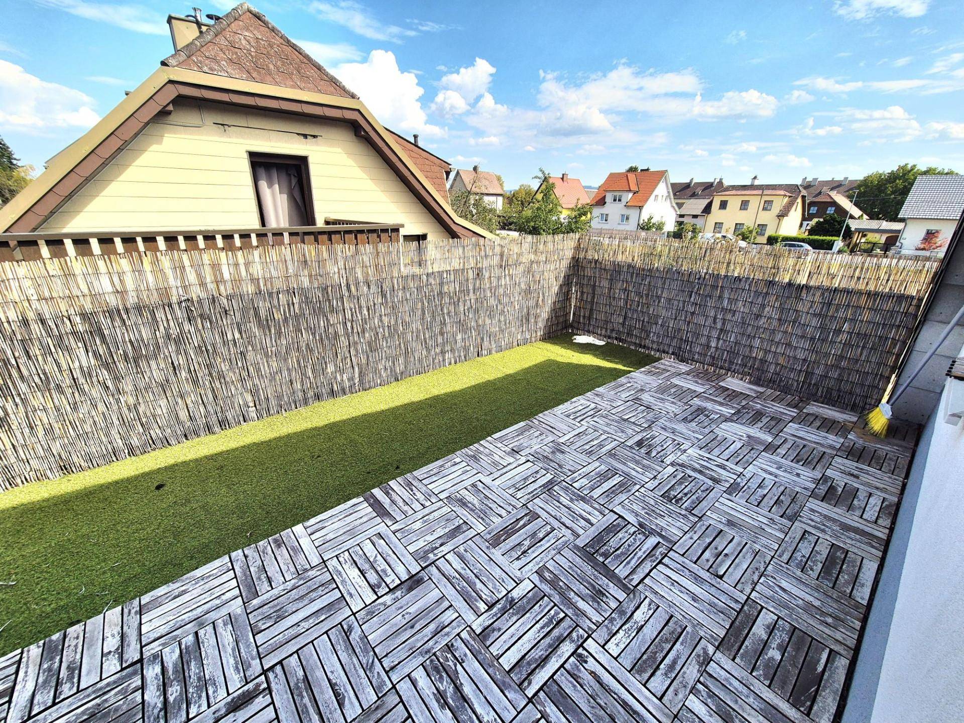 Kleiner privater Gartenbereich mit Holzfliesen und Kunstrasen, umgeben von einem Sichtschutzzaun.