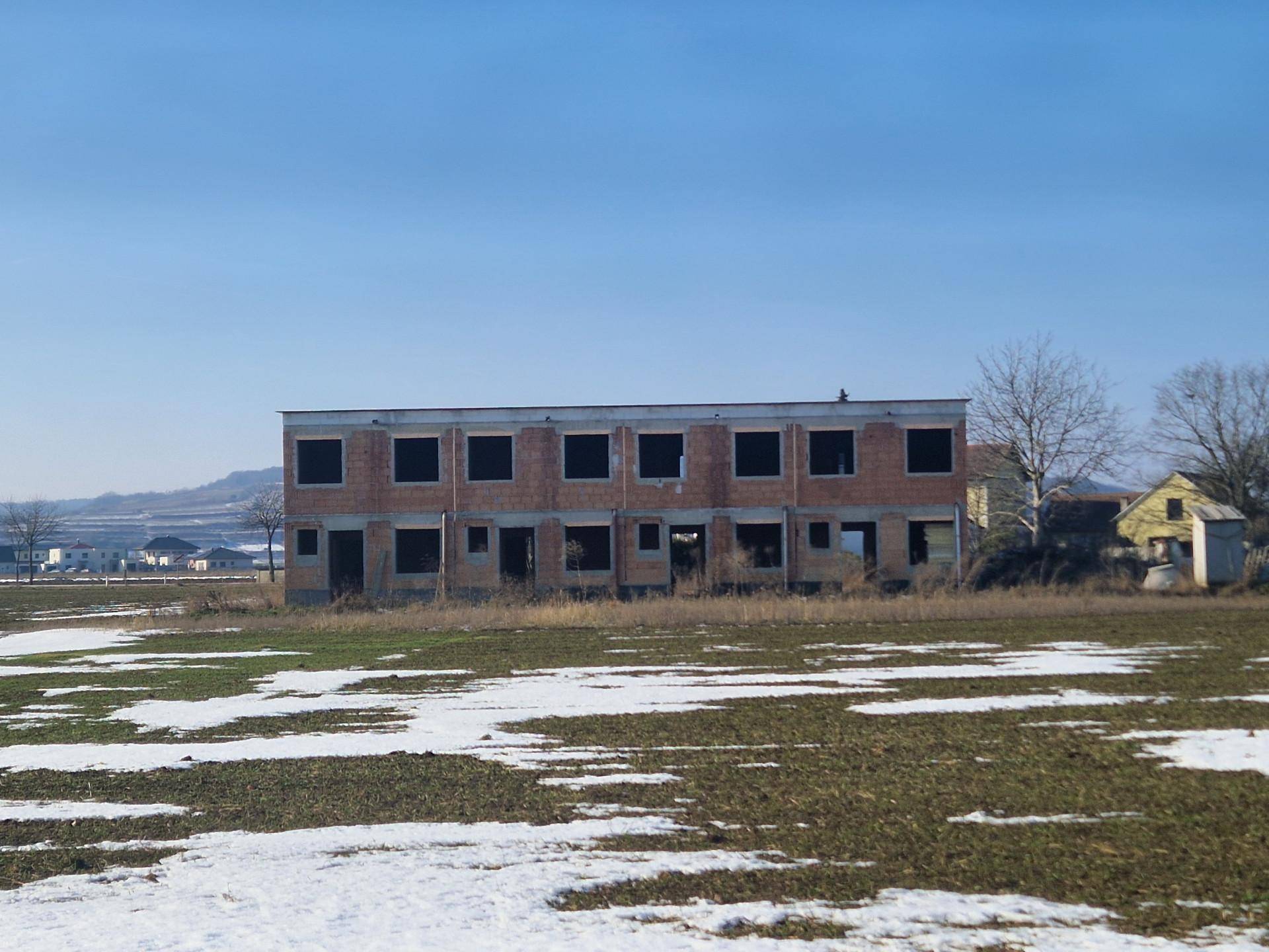 Unfertiges Gebäude im Rohbauzustand mit offenen Fensteröffnungen und Schneeresten auf dem Feld im Winter.