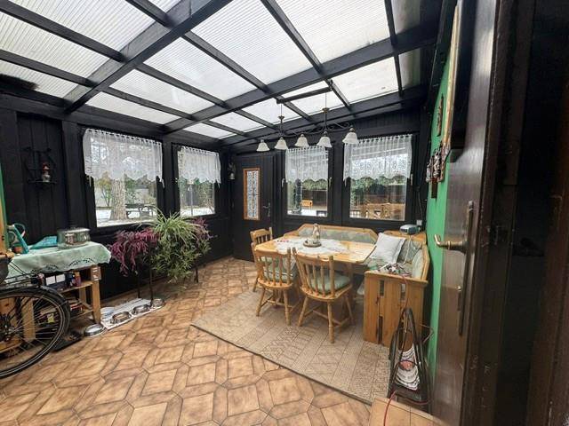 Gemütlicher Wintergarten mit Holztisch, Eckbank und Fliesenboden, umgeben von großen Fenstern.