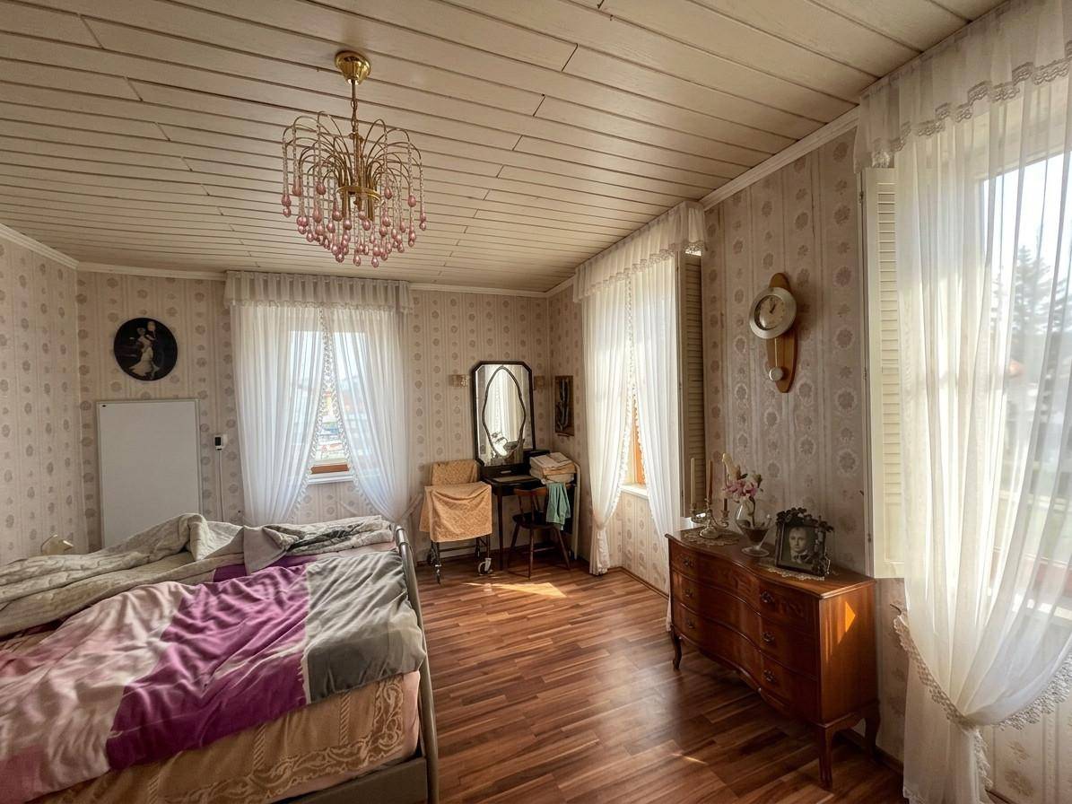 Schlafzimmer mit gemusterter Tapete, Holzdecke und zwei Fenstern, die viel Tageslicht hereinlassen.