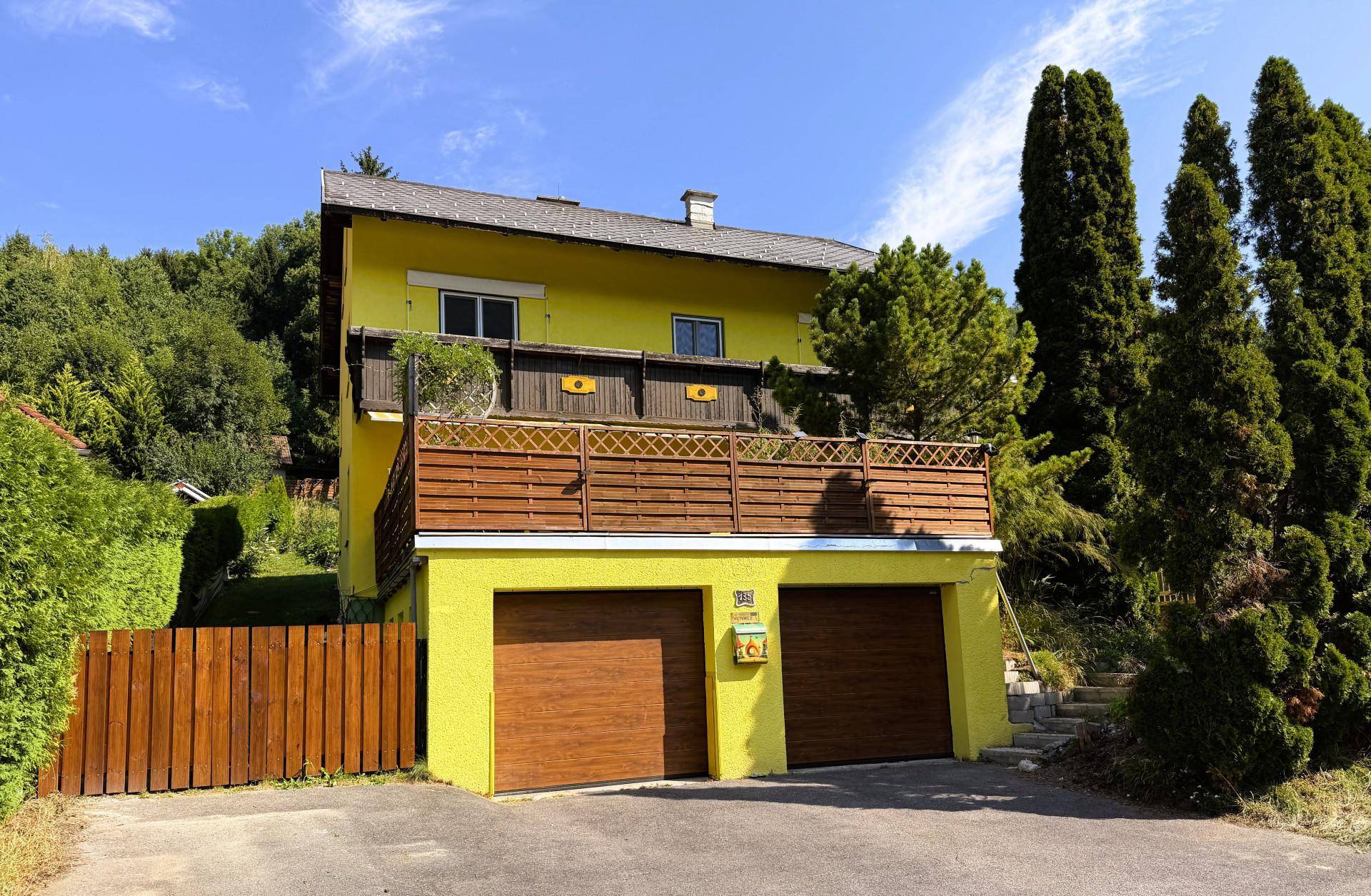 Gelbes Haus mit zwei Garagen und einem Balkon, umgeben von hohen Bäumen und grüner Natur.
