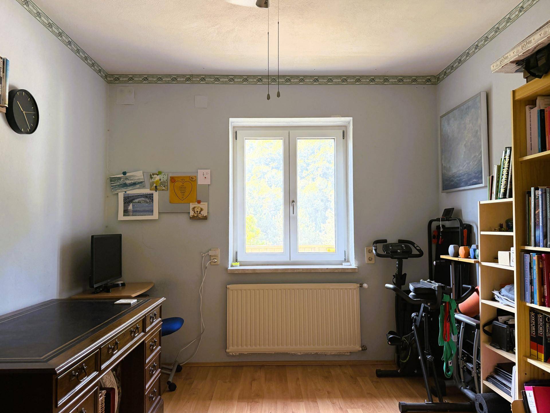 Arbeitszimmer mit Holzboden, großem Schreibtisch, Bücherregal und Fenster mit Blick ins Grüne.