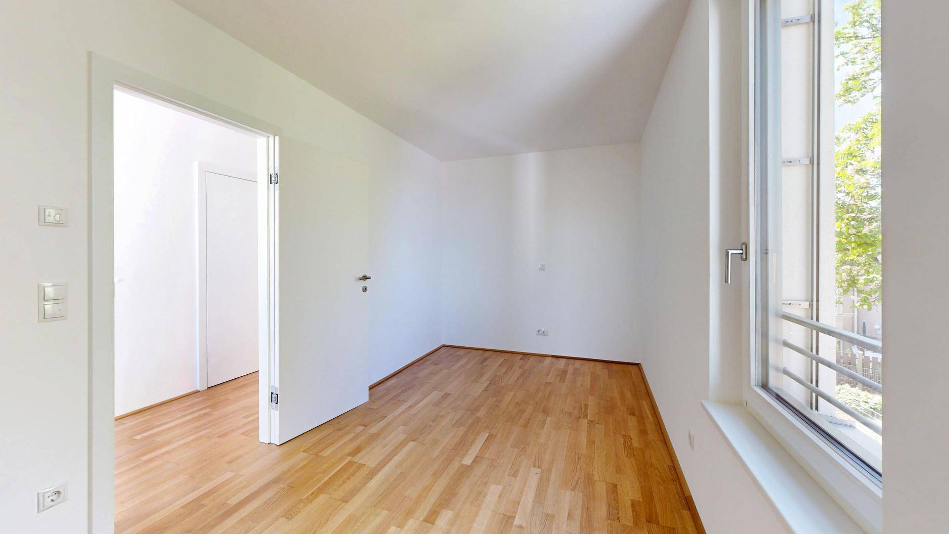 Leeres Zimmer mit Parkettboden und großem Fenster, das einen grünen Ausblick bietet.