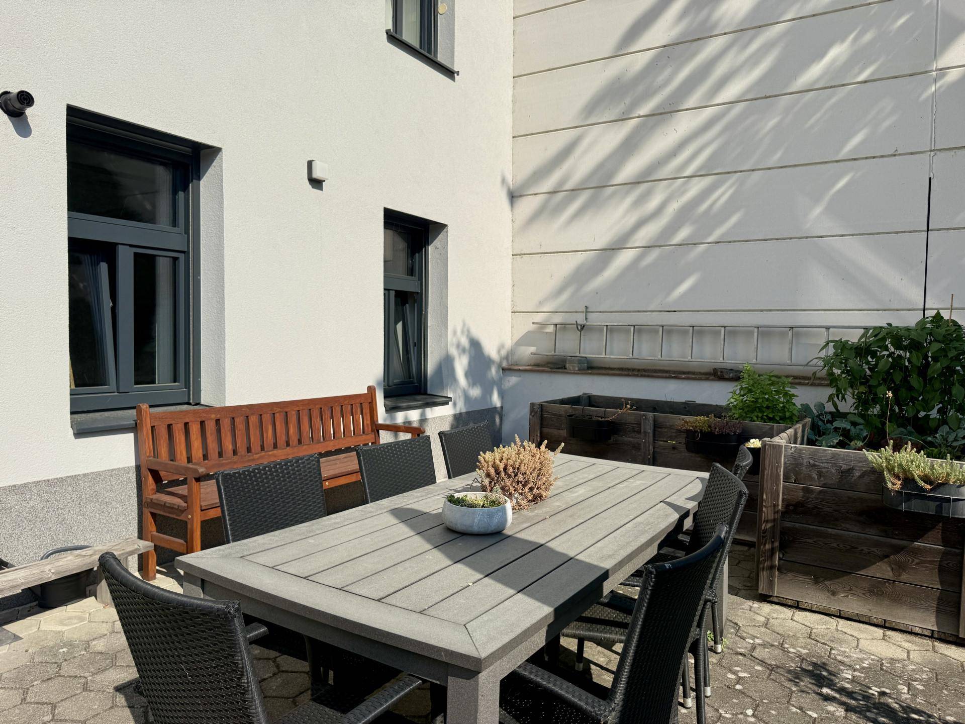 Sonniger Außenbereich mit Esstisch, Stühlen und Hochbeeten an der Hauswand, ideal zum Entspannen.