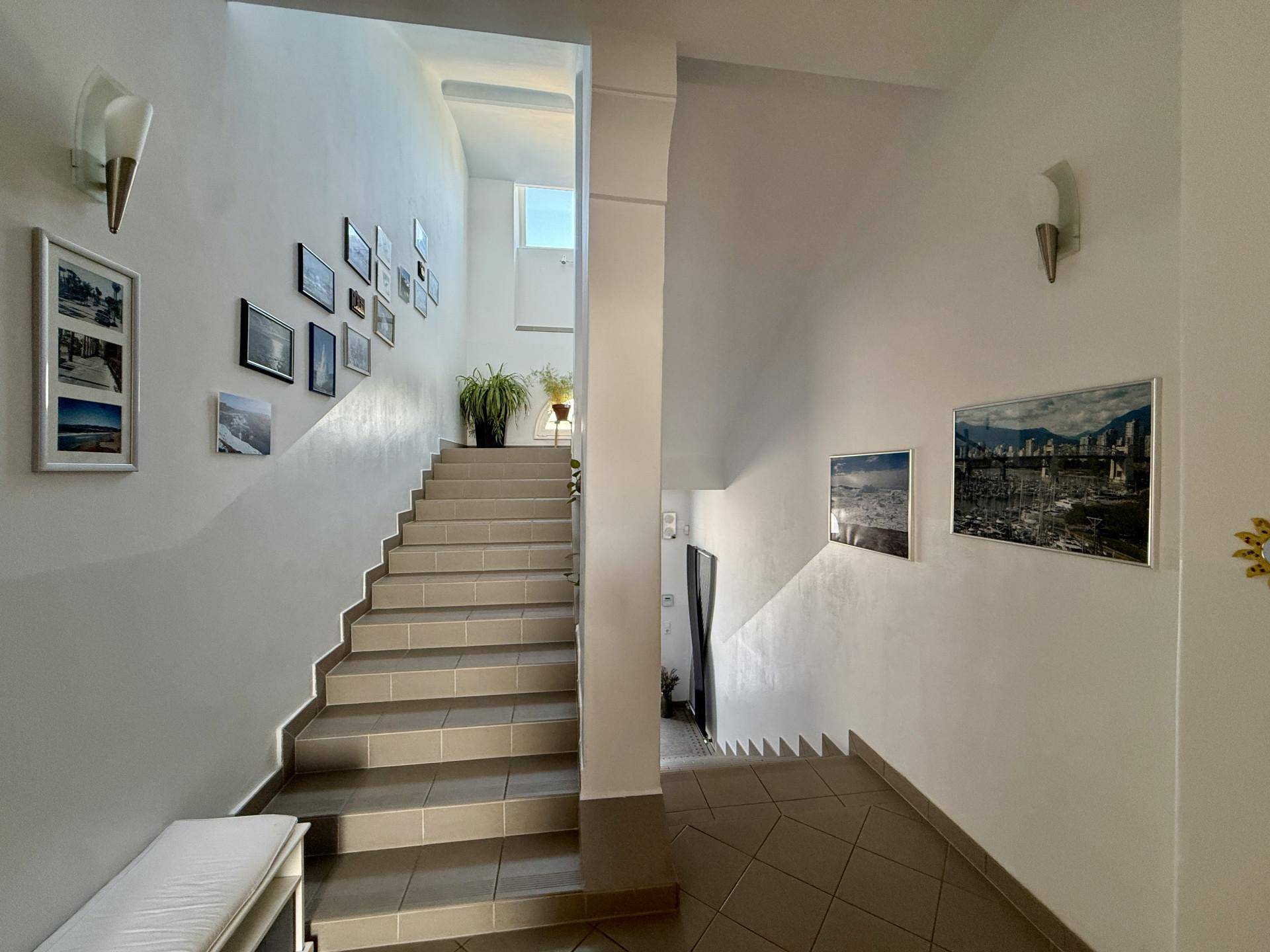 Helles Treppenhaus mit Fliesenboden und gerahmten Bildern an den Wänden, lichtdurchflutet.