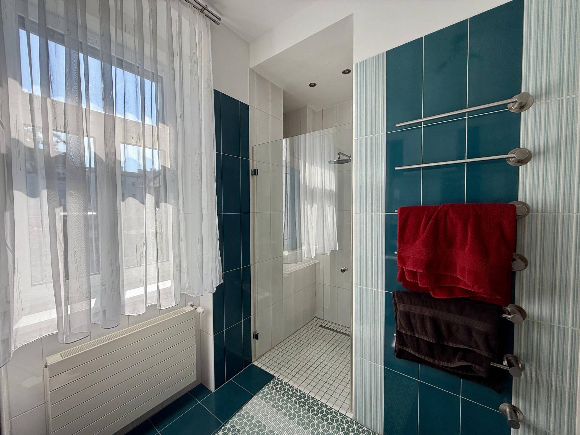 Helles Badezimmer mit begehbarer Dusche, blauen Fliesen und beheiztem Handtuchhalter am Fenster.