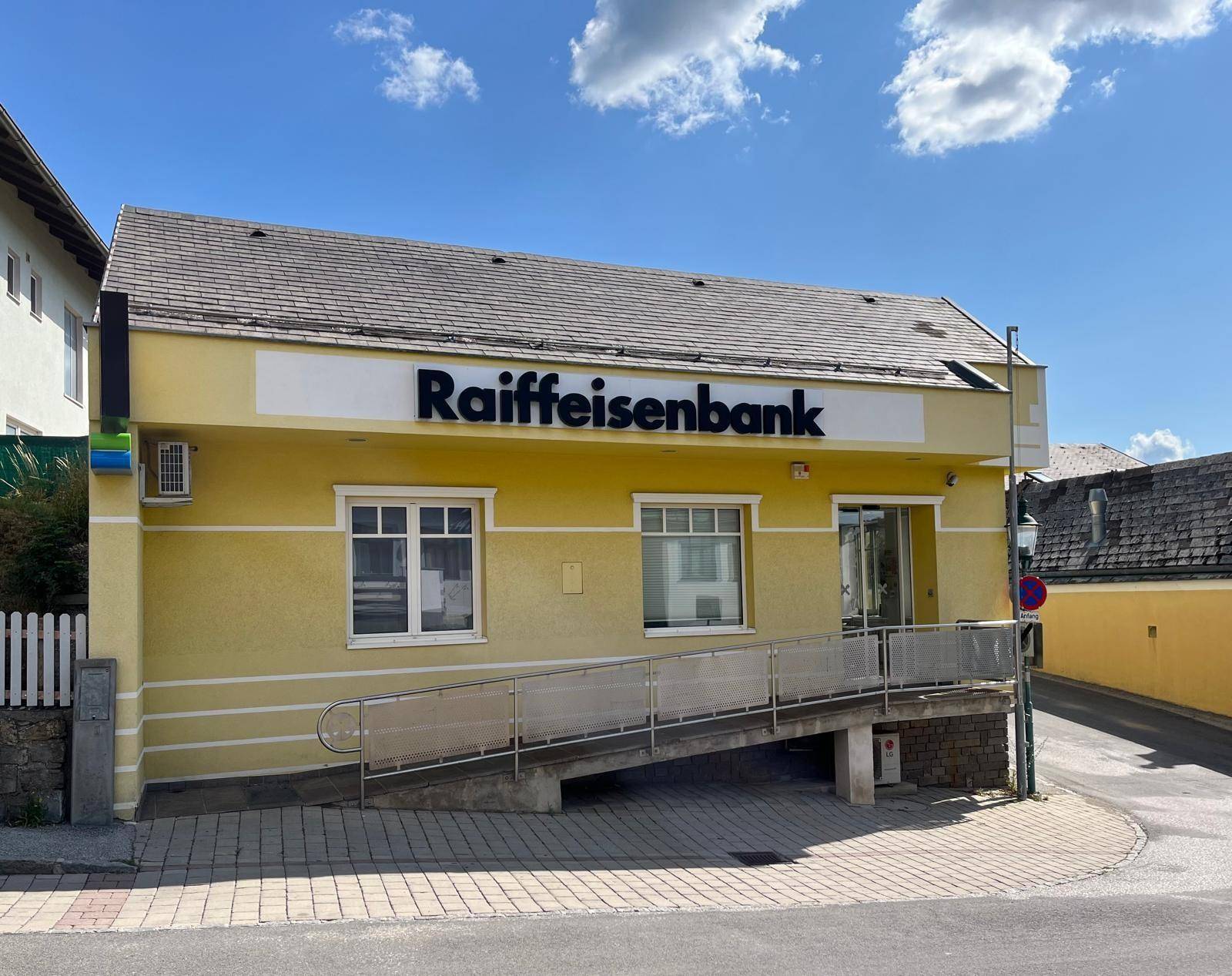 Ansicht des gelben Bankgebäudes mit Rampe und Raiffeisenbank-Logo an einem sonnigen Tag.