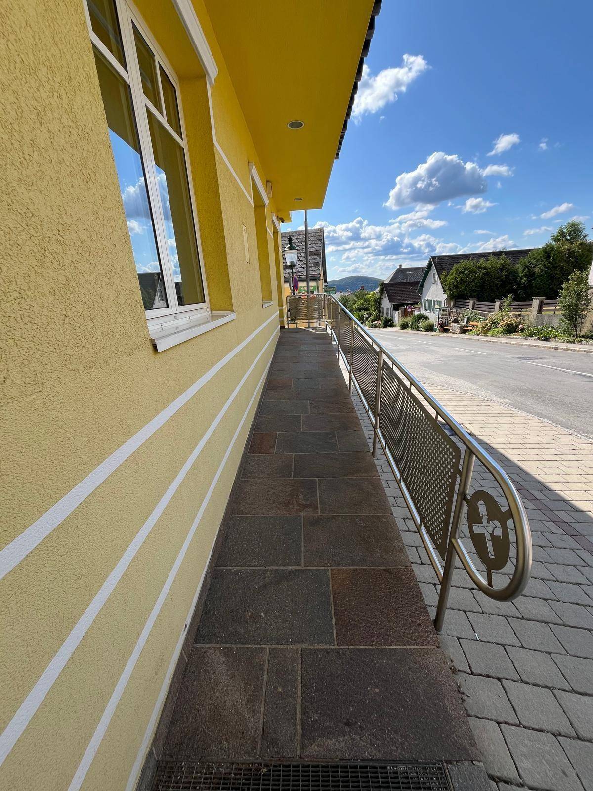 Barrierefreie Rampe mit Metallgeländer und Steinplattenboden vor dem gelben Gebäude.