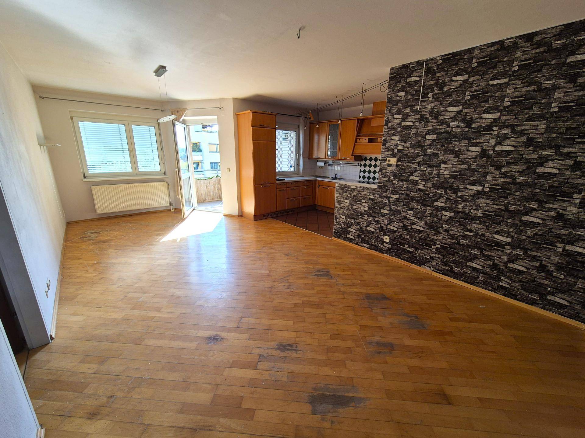 Geräumiger Wohnbereich mit Holzboden, Zugang zum Balkon und integrierter Küche im Hintergrund.