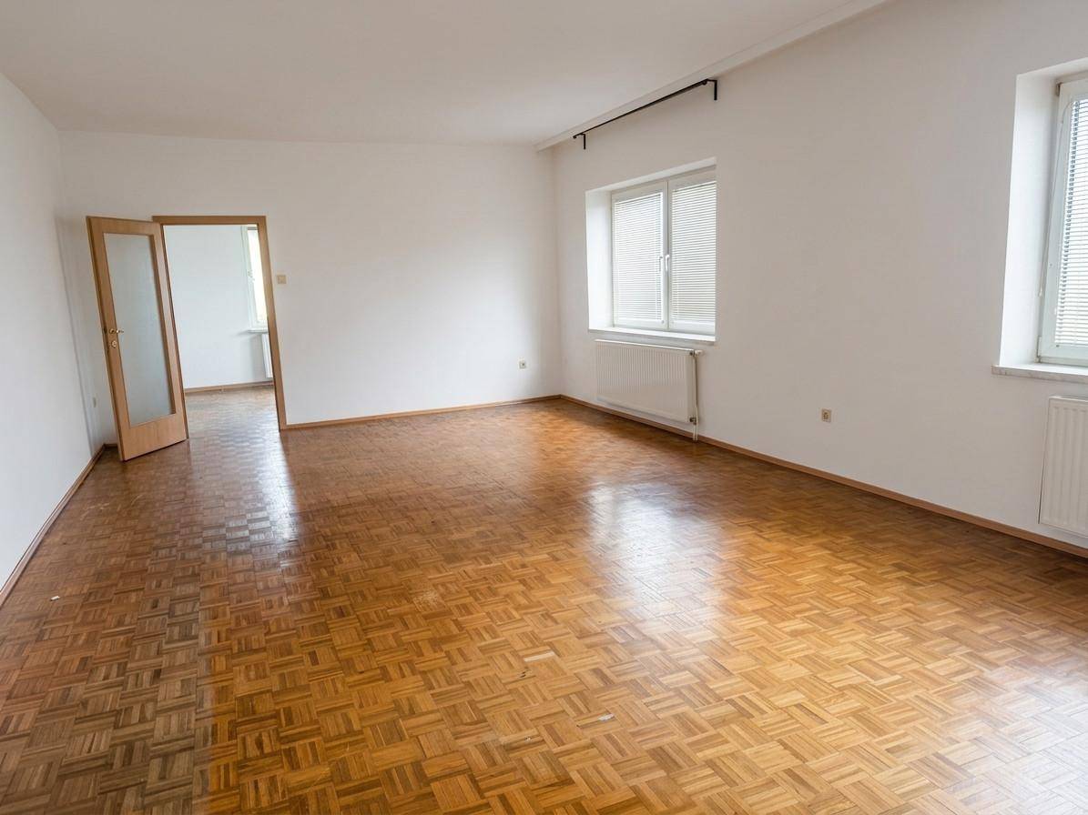 Helles, leeres Zimmer mit Parkettboden und zwei Fenstern, ideal als Wohn- oder Schlafzimmer.