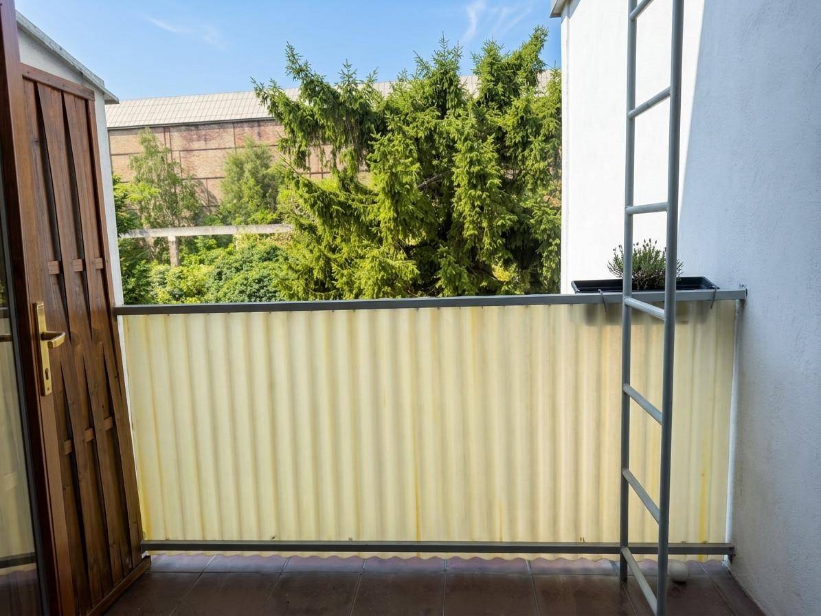 Kleiner Balkon mit Sichtschutz und Blick auf Bäume sowie ein entferntes Industriegebäude.