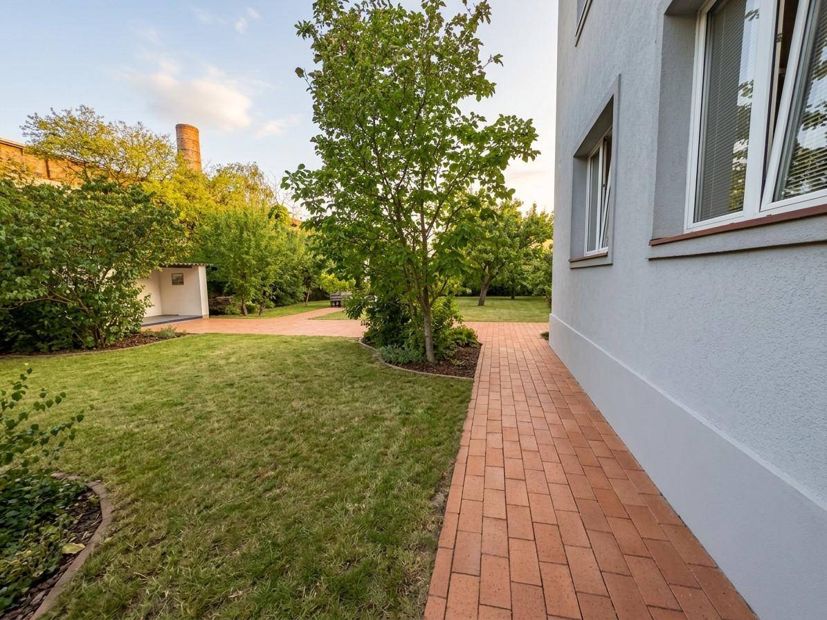Gepflasterter Weg entlang des Hauses führt zu einem grünen Gartenbereich mit Rasen und Bäumen.