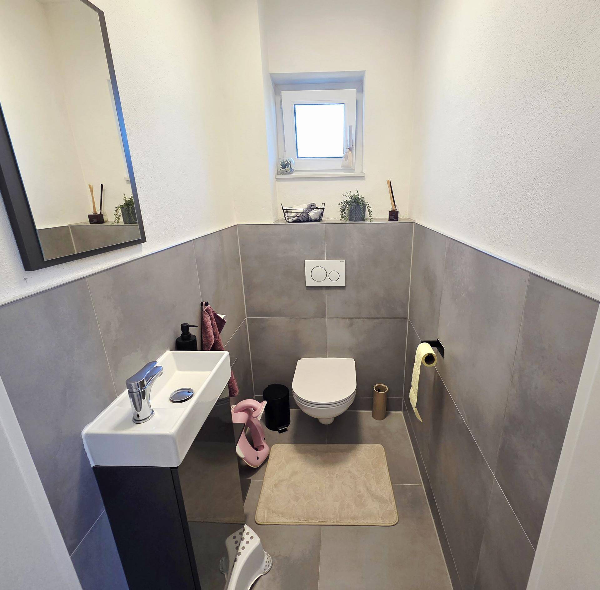 Modernes Gäste-WC mit wandhängender Toilette, kleinem Waschbecken und grauen Fliesen.
