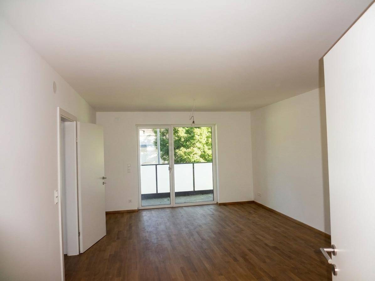 Leeres Zimmer mit Holzboden und großem Schiebefenster, das Zugang zu einem Balkon bietet.