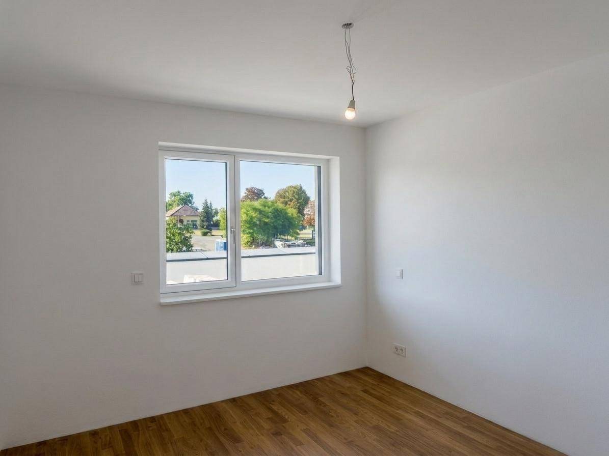 Helles, leeres Zimmer mit Holzboden und einem Fenster, das einen Blick ins Grüne bietet.