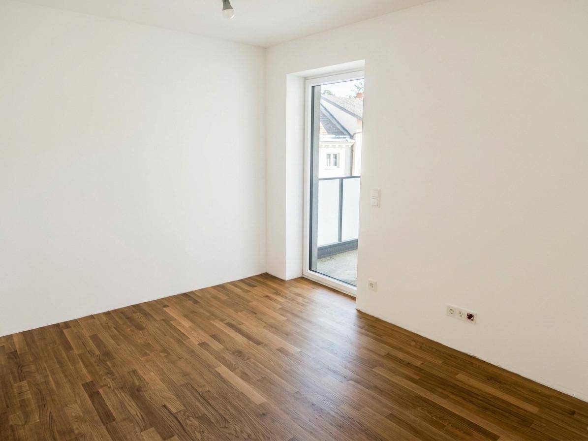 Leeres Zimmer mit Holzboden und Zugang zu einem kleinen Balkon oder einer französischen Balkontür.