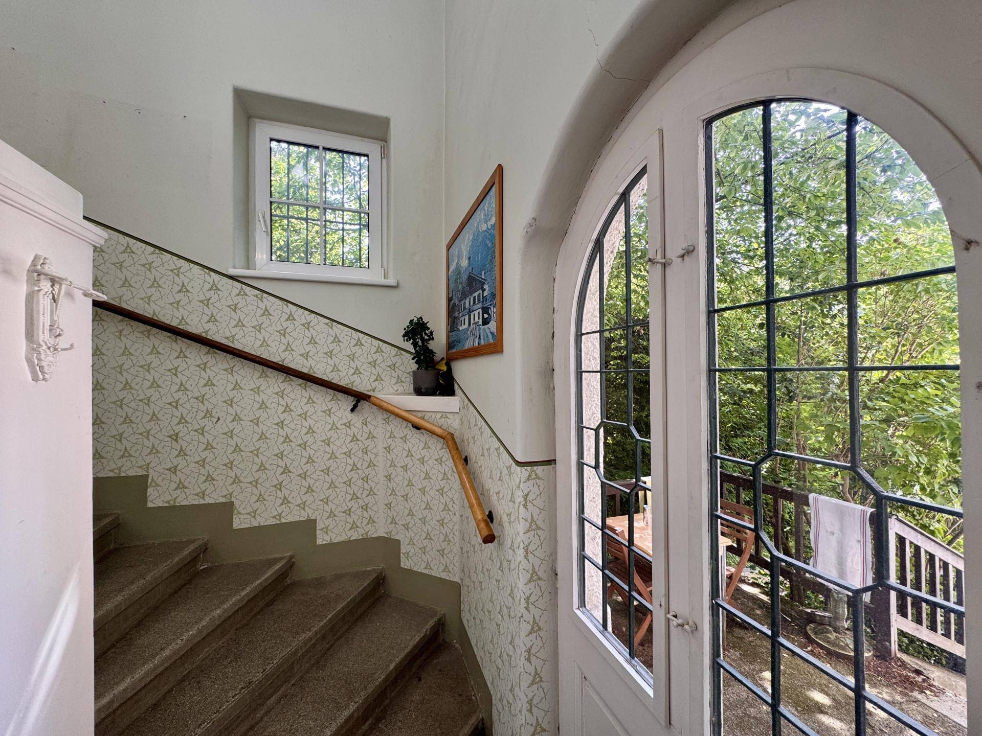 Treppenhaus mit gemusterter Tapete, Fenster und einer verglasten Tür zum Außenbereich mit Bäumen.