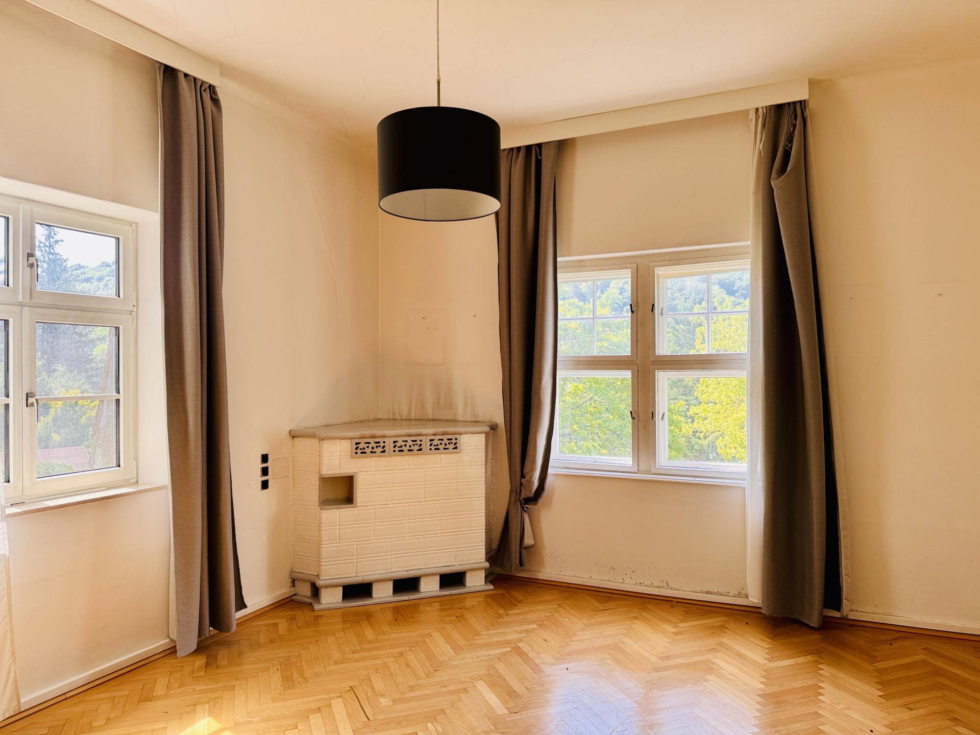 Leeres Zimmer mit Fischgrätparkett, zwei Fenstern und einem traditionellen Kachelofen in der Ecke.