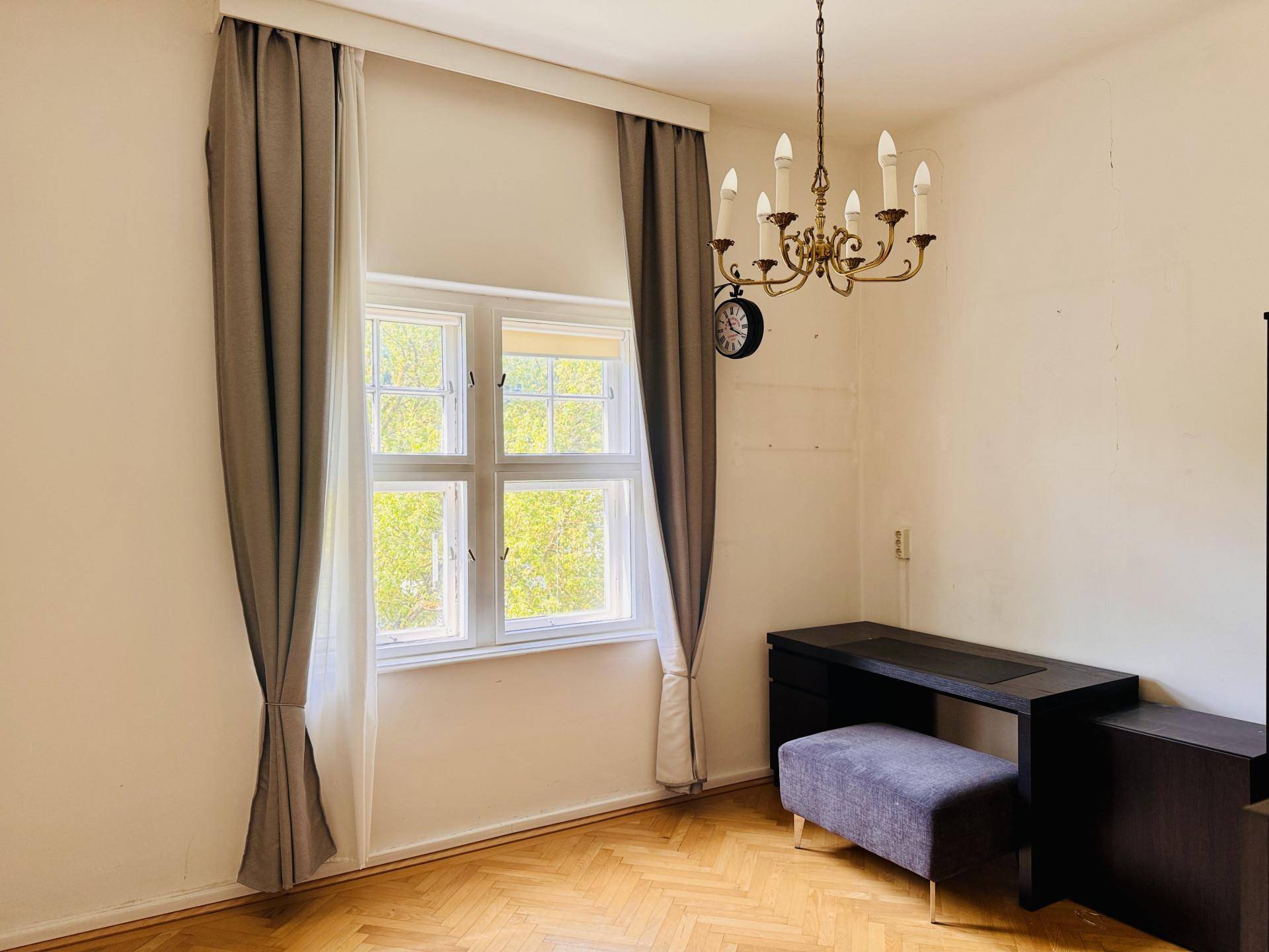 Helles Zimmer mit Fischgrätparkett, Kronleuchter und einem dunklen Schreibtisch unter dem Fenster.