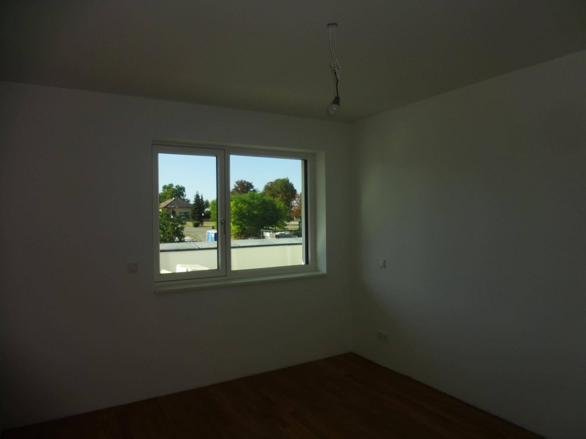 Unmöbliertes Zimmer mit Holzboden und Fenster mit Blick ins Grüne.