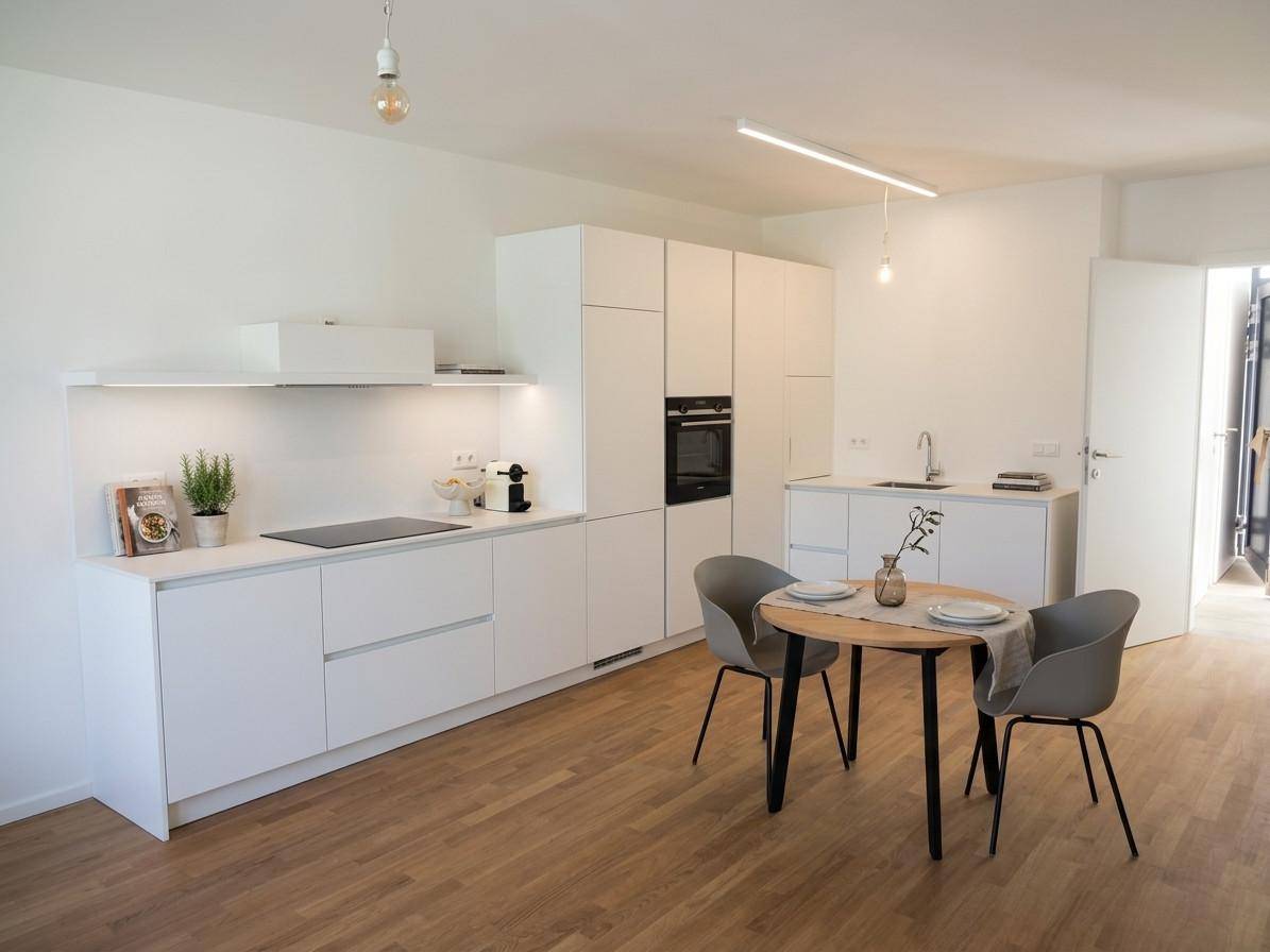 Moderne weiße Küche mit Holzboden, integrierten Geräten und kleinem Essbereich.