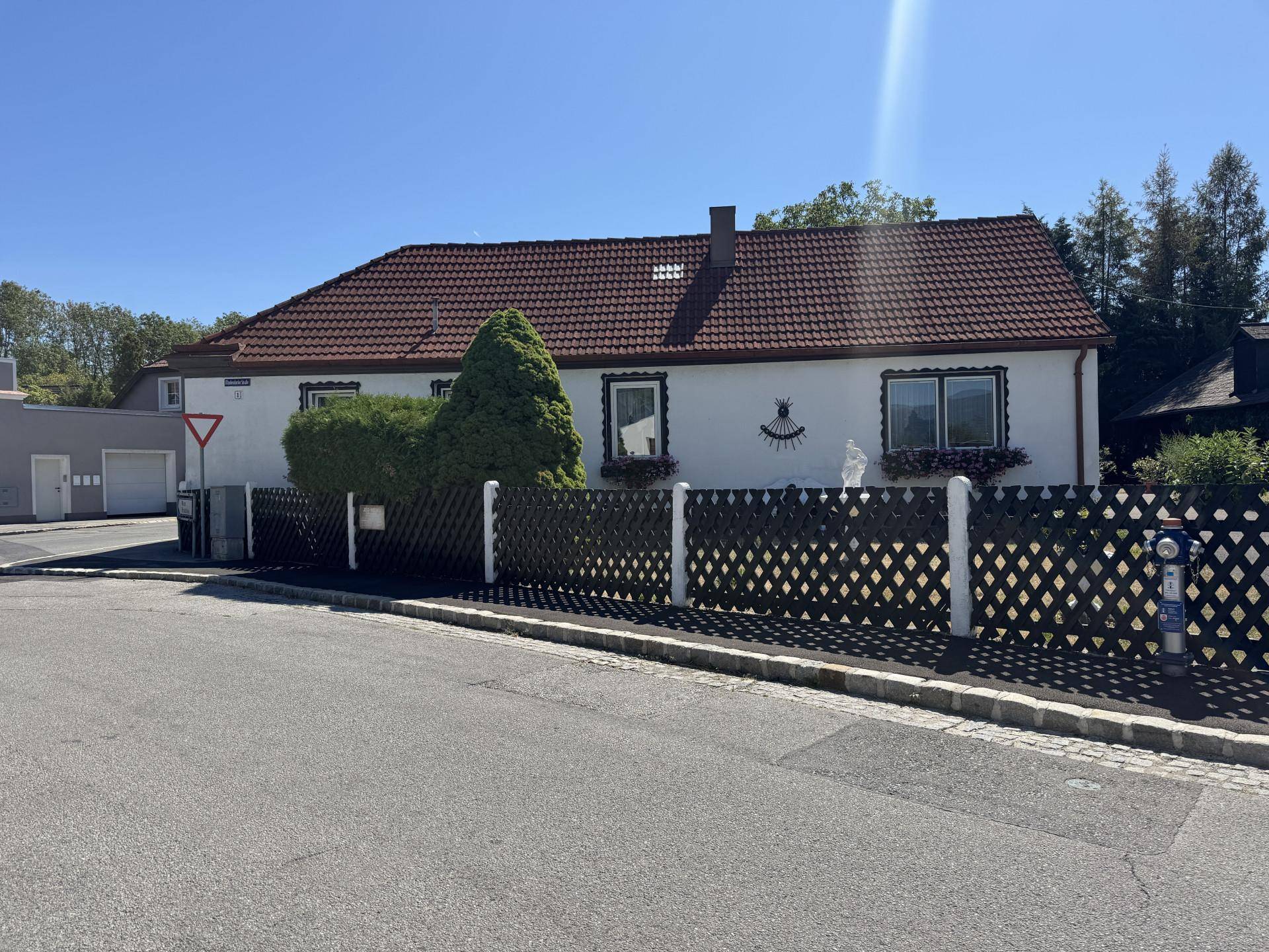 Hausfassade von der Straße aus gesehen mit Zaun, Fenstern und rotem Ziegeldach.