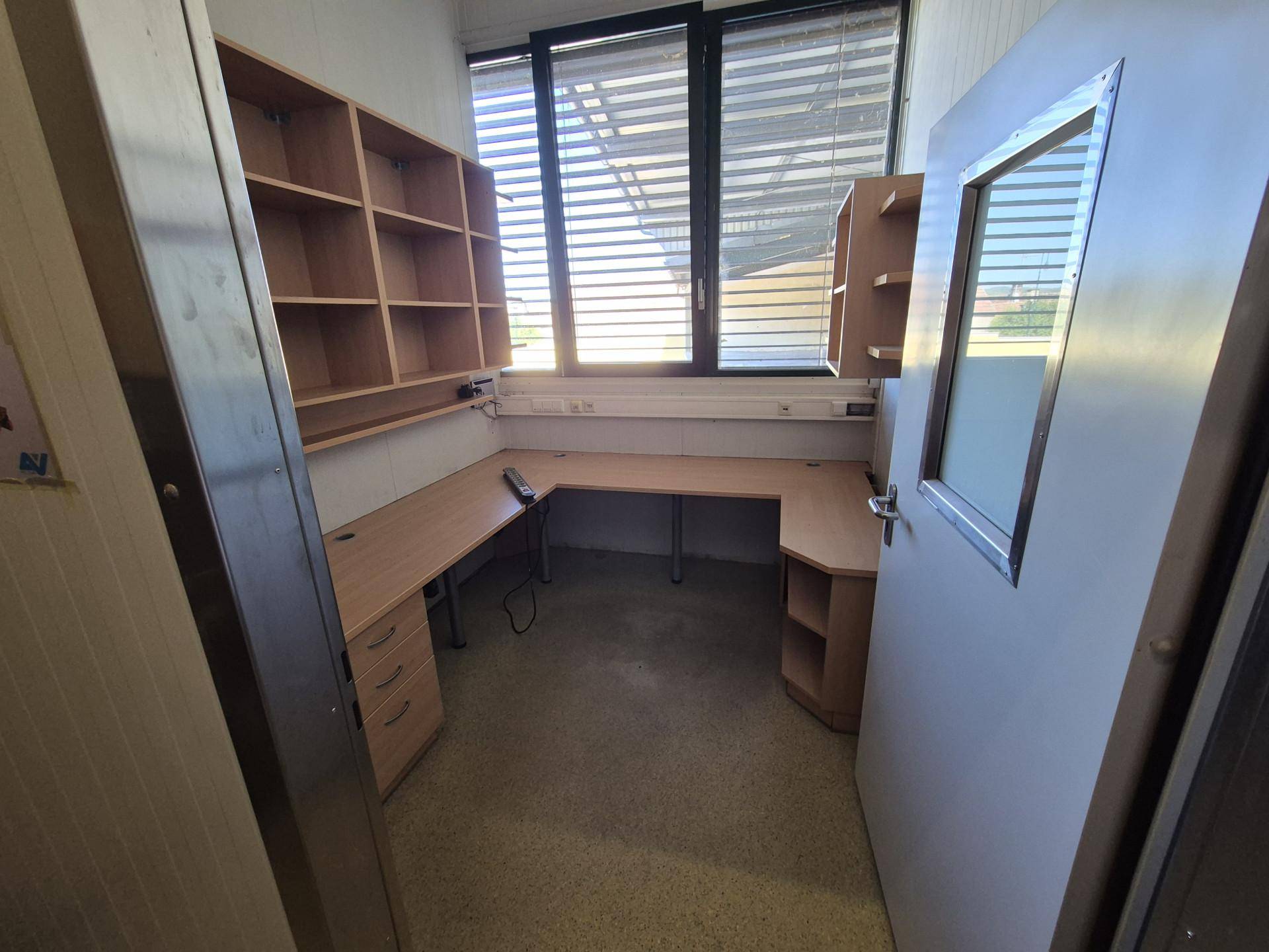 Büroraum mit Schreibtisch, Regalen und einem großen Fenster mit Jalousien.