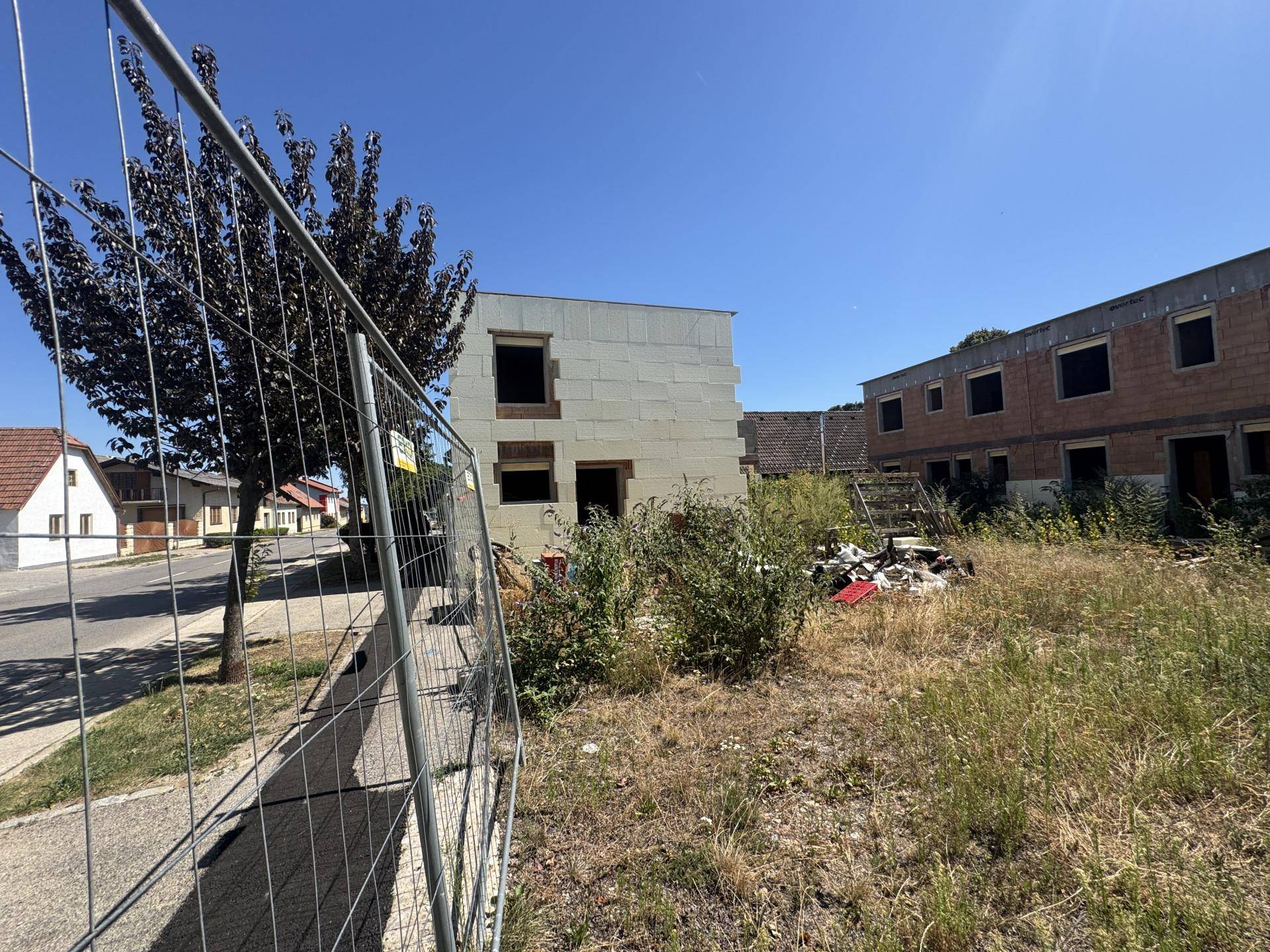 Unfertiges Gebäude hinter einem Bauzaun mit überwuchertem Gelände und weiteren Häusern im Hintergrund.