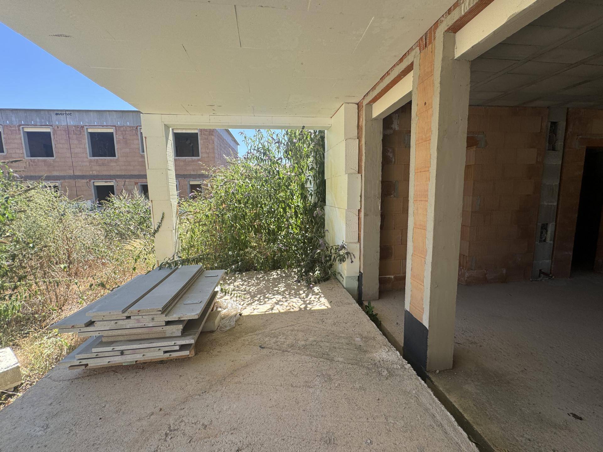Unfertiger Innenraum mit Betonboden und Blick auf die überwucherte Außenanlage.