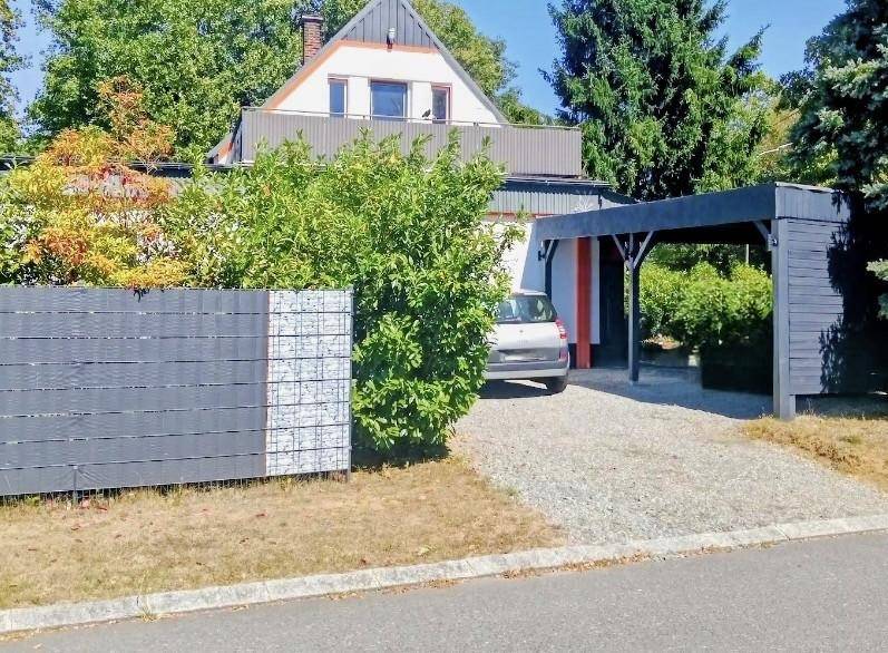 Vorderansicht des Hauses mit Carport, Einfahrt und umgebender Bepflanzung bei Sonnenschein.