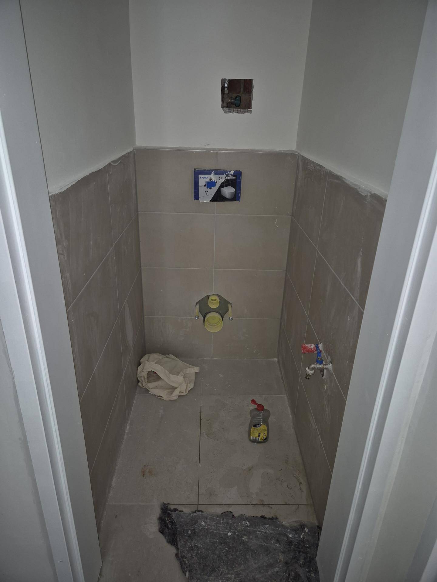 Kleiner, unfertiger Raum mit grauen Fliesen und vorbereiteten Anschlüssen für eine Toilette.