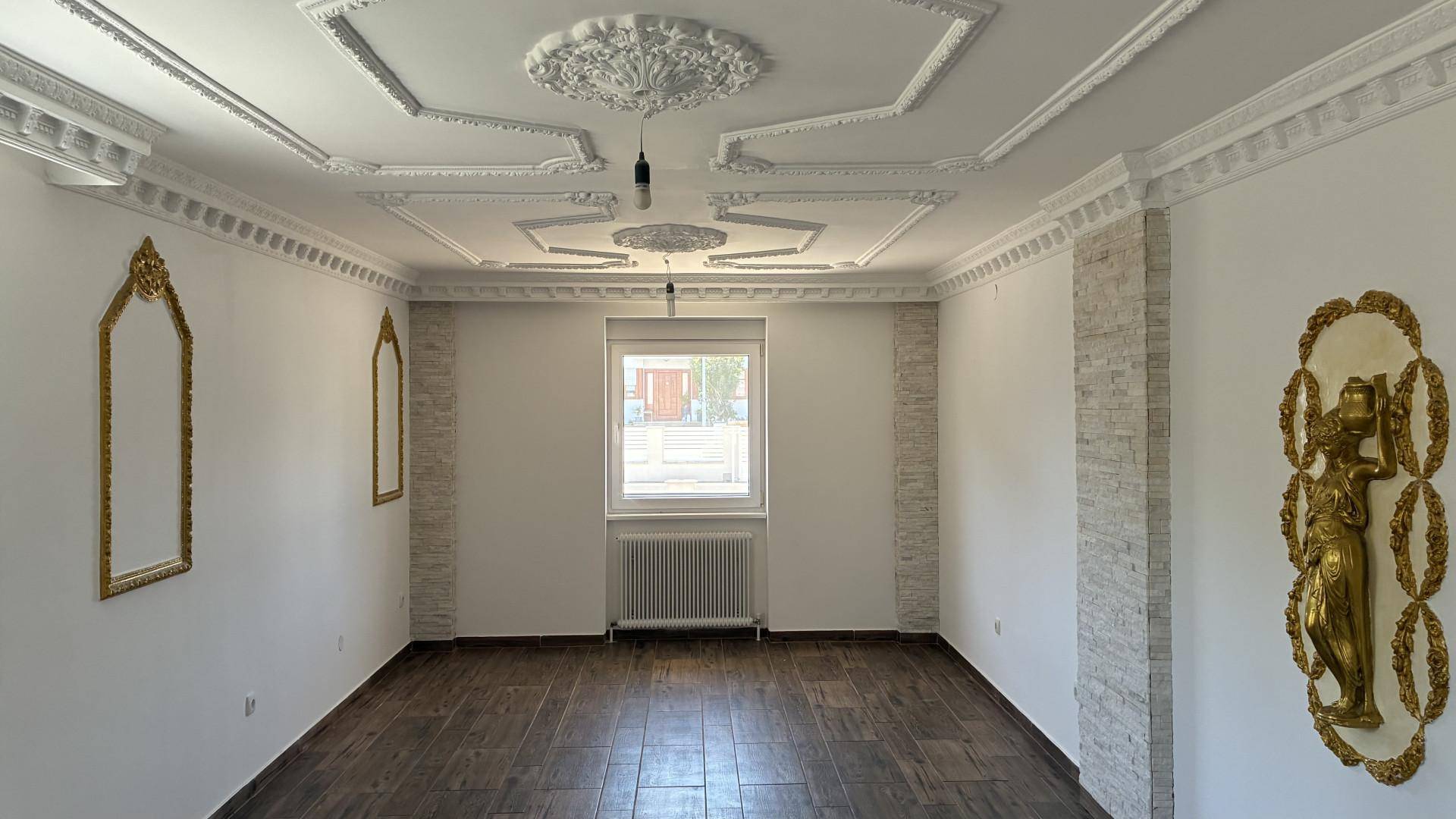Geräumiges Wohnzimmer mit aufwendiger Stuckdecke, großem Fenster und dekorativen Wandelementen.