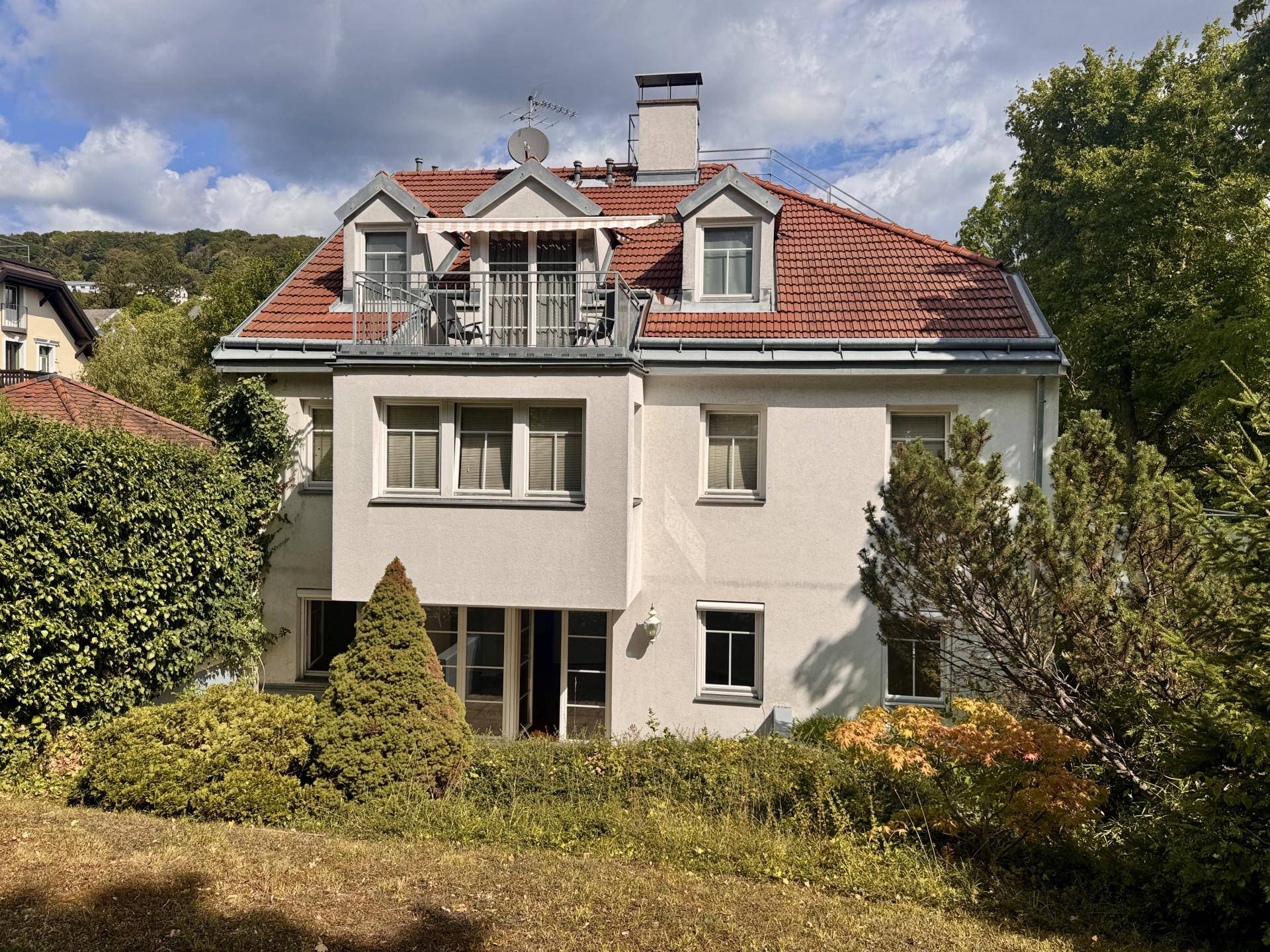 Charmantes Haus mit rotem Ziegeldach, Balkon und grüner Umgebung unter bewölktem Himmel.