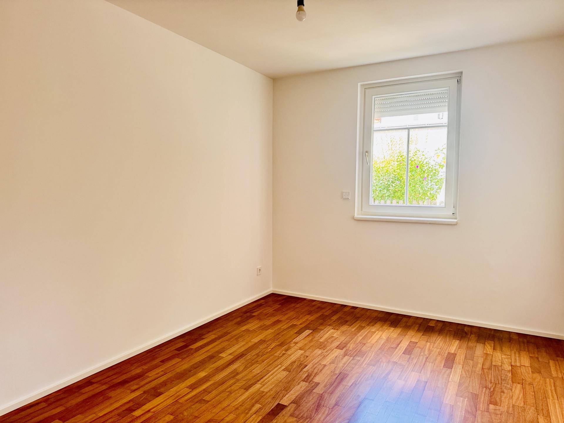Freundliches Zimmer mit Parkettboden und Fenster, das viel Tageslicht hereinlässt.