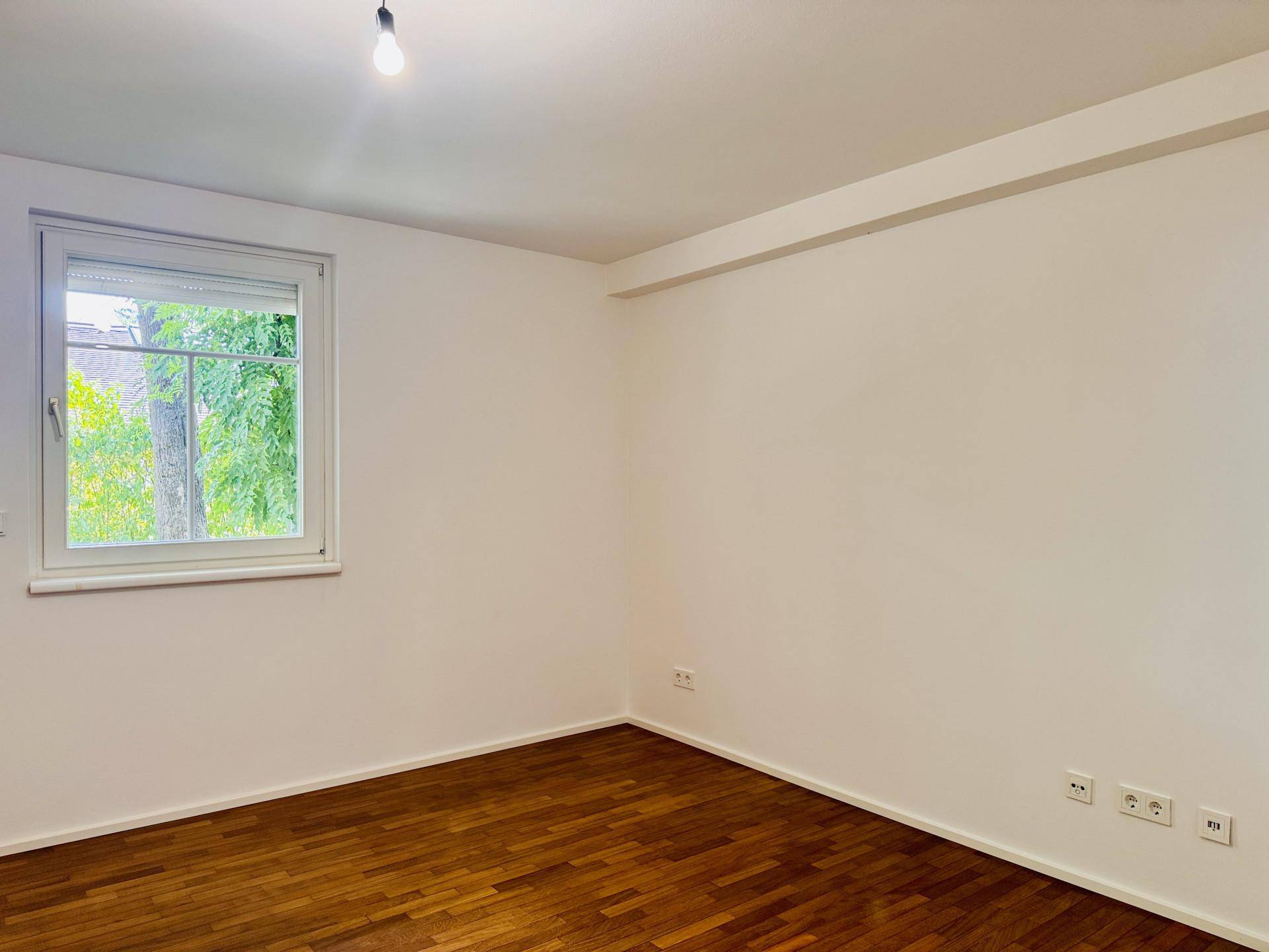 Helles Zimmer mit Parkettboden und Fenster, das einen Blick ins Grüne bietet.